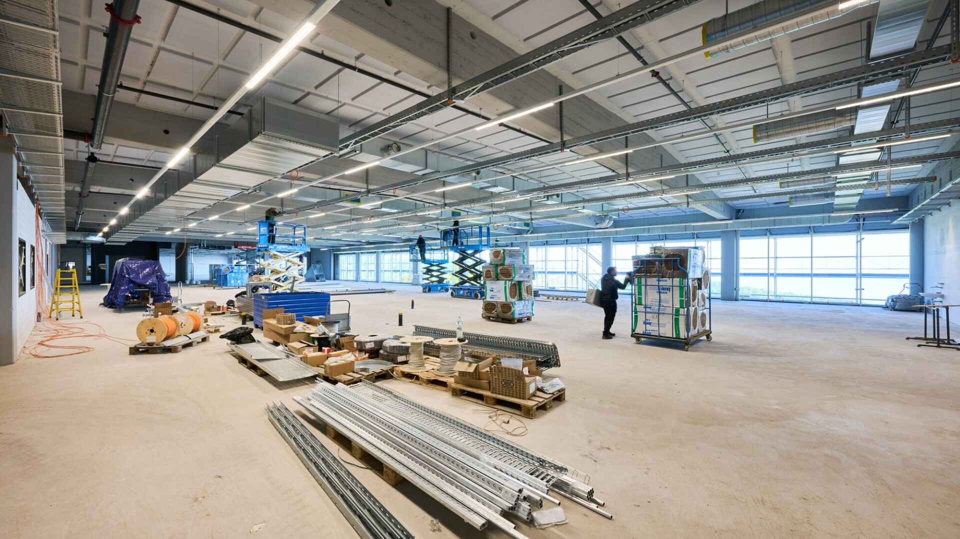 Große, helle Industriehalle im Bau. Verstreute Baumaterialien, Metallträger, Geräte und ein Arbeiter bewegen eine Palette. Sichtbare Deckenplatten und große Fenster sorgen für Tageslicht.