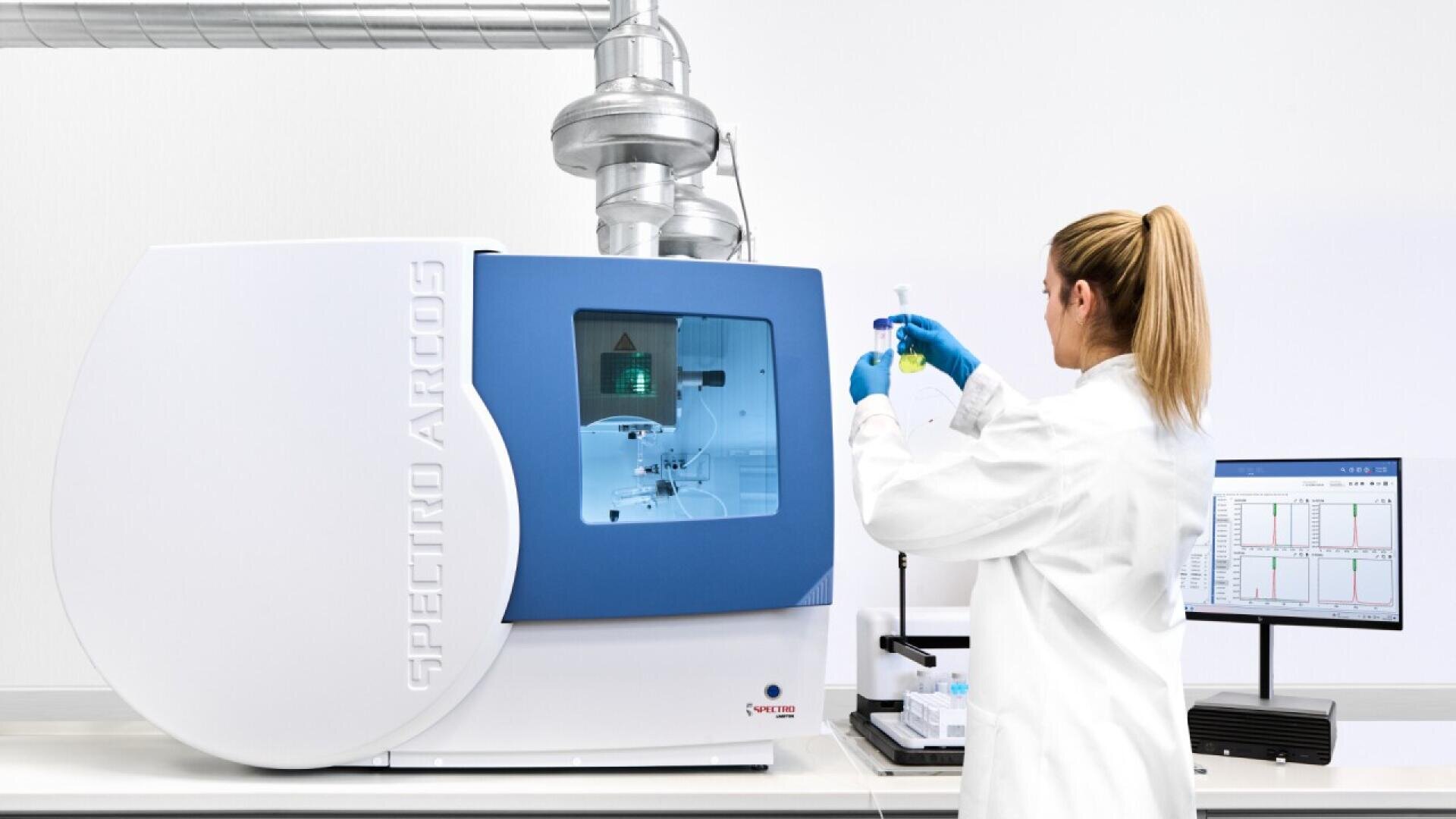 Un scientifique en blouse blanche tient une fiole contenant un liquide vert, à côté d'un instrument d'analyse SPECTRO ARCOS dans un laboratoire moderne. Un ordinateur avec des graphiques se trouve sur le bureau à proximité.