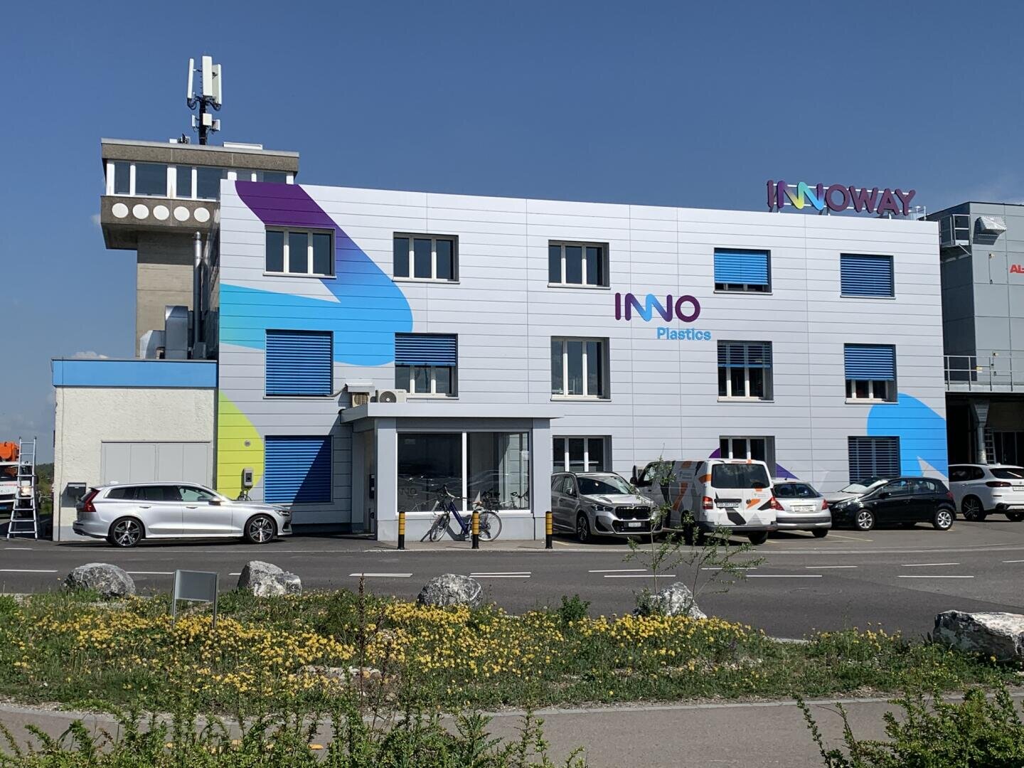 Ein modernes Bürogebäude mit der Aufschrift INNO Plastics mit bunten geometrischen Akzenten, parkenden Autos davor und einem Schild mit der Aufschrift INNOWAY auf dem Dach vor einem klaren blauen Himmel.