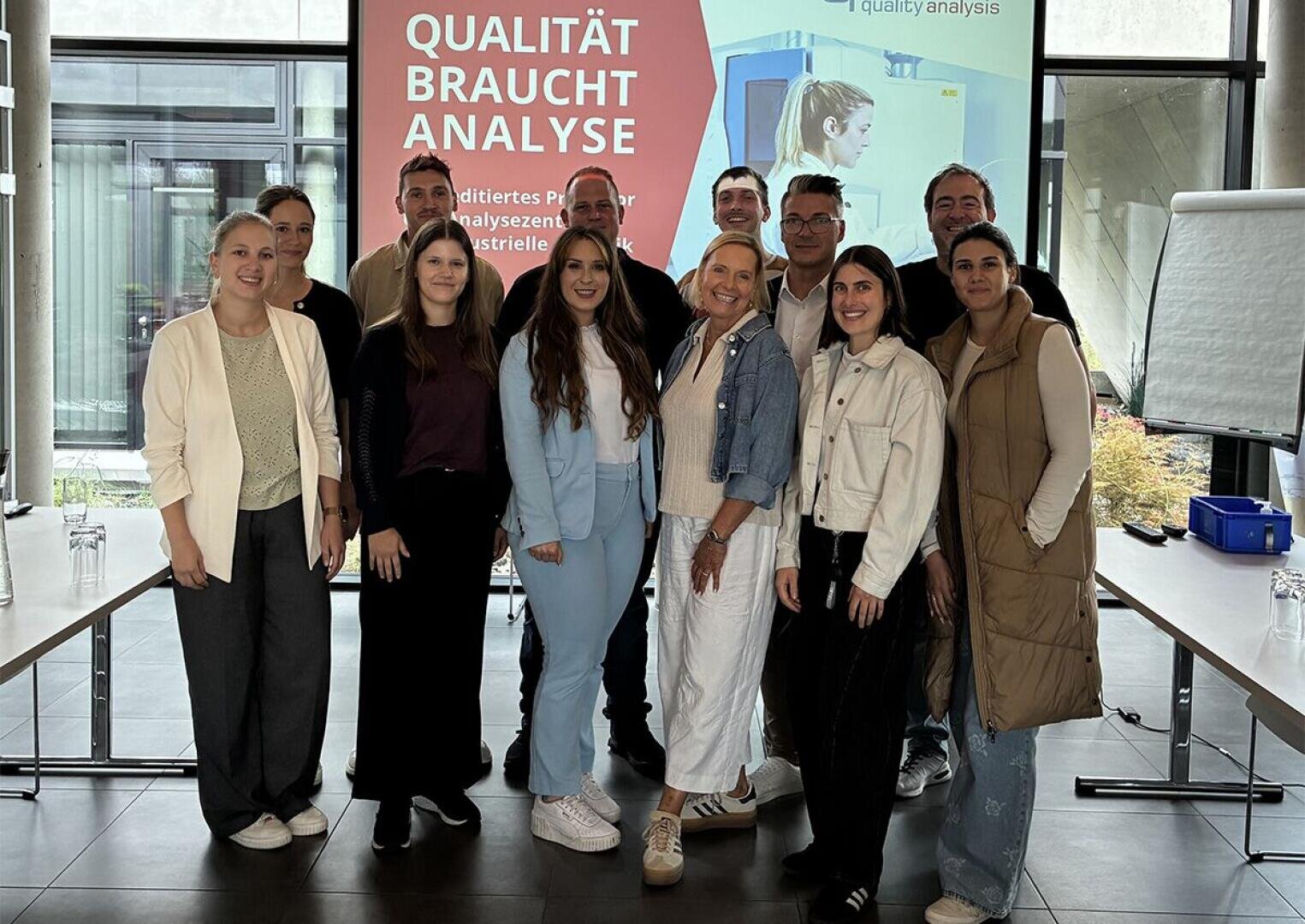 Eine Gruppe von dreizehn Personen steht lächelnd in einem Raum vor einem Bildschirm mit der Aufschrift QUALITÄT BRAUCHT ANALYSE. Sie scheinen an einer professionellen Veranstaltung oder einem Workshop teilzunehmen.