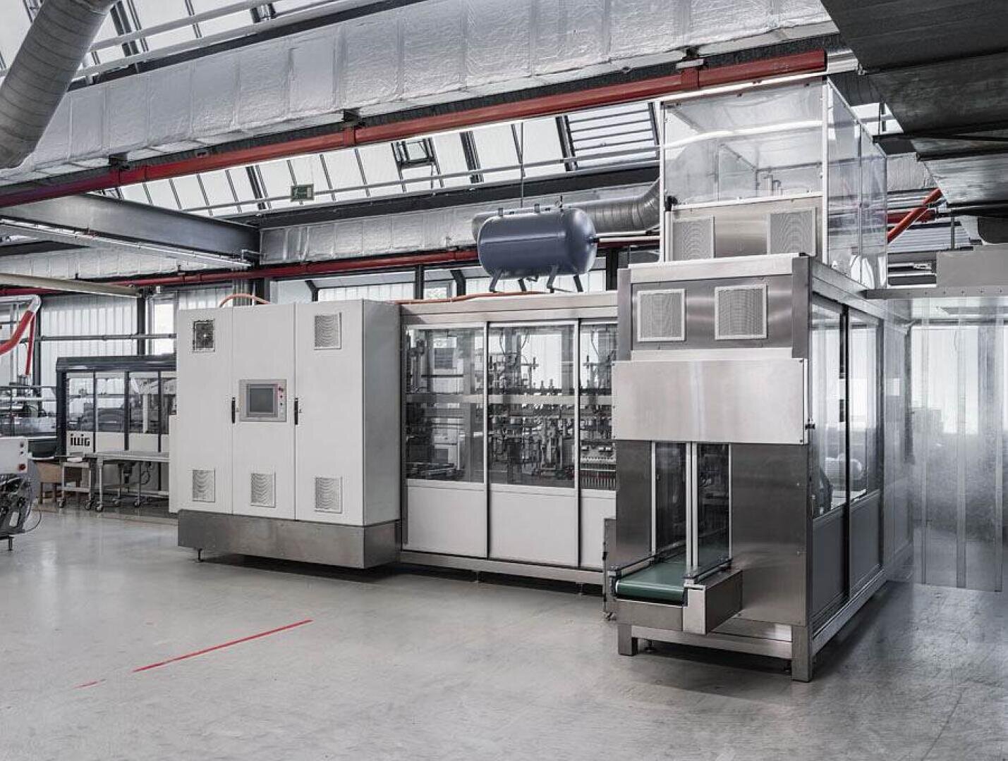 Eine moderne Industriemaschine in einer geräumigen, gut beleuchteten Fabrikhalle mit Edelstahltafeln, einem Kontrollbildschirm und einem Förderband unter transparenten Schutzabdeckungen.