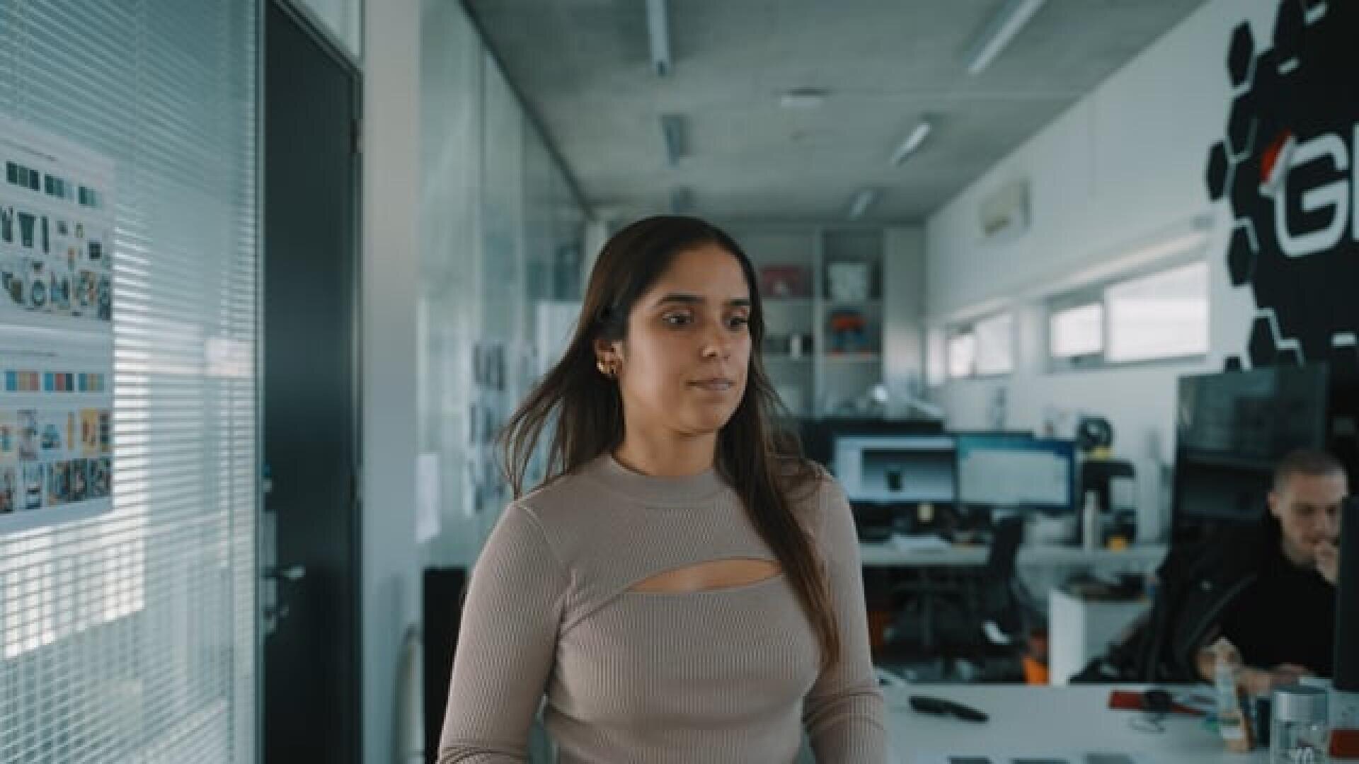Une femme aux longs cheveux bruns portant un haut beige avec une découpe se tient dans un bureau moderne avec des bureaux, des ordinateurs et une autre personne travaillant à l'arrière-plan.