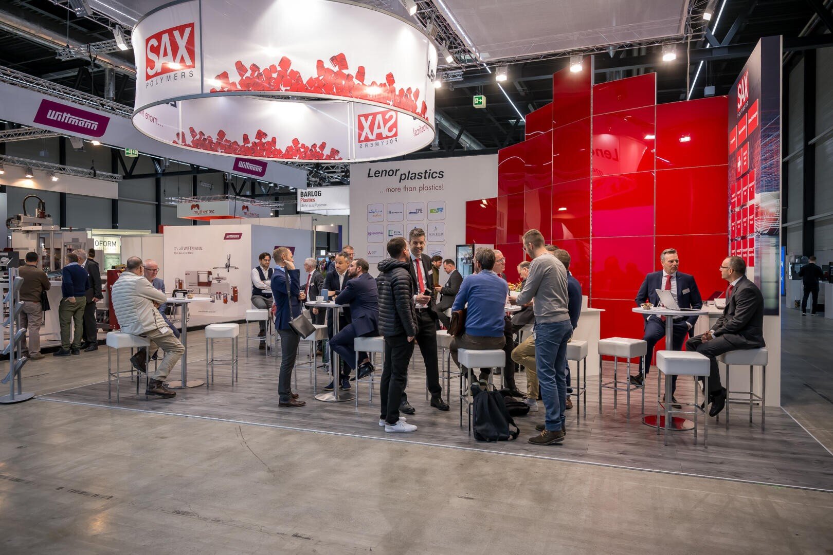 Eine Gruppe von Menschen interagiert und knüpft Kontakte an einem Messe- oder Konferenzstand mit rot-weißer Beschilderung, modernen Möbeln und Informationsdisplays auf einem polierten Betonboden.