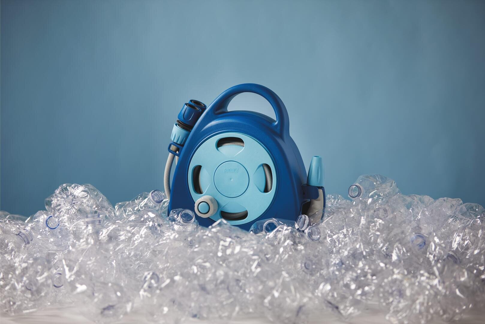 Eine blaue Gartenschlauchtrommel ist umgeben von einem Haufen zerknitterter klarer Plastikflaschen vor einem hellblauen Hintergrund.