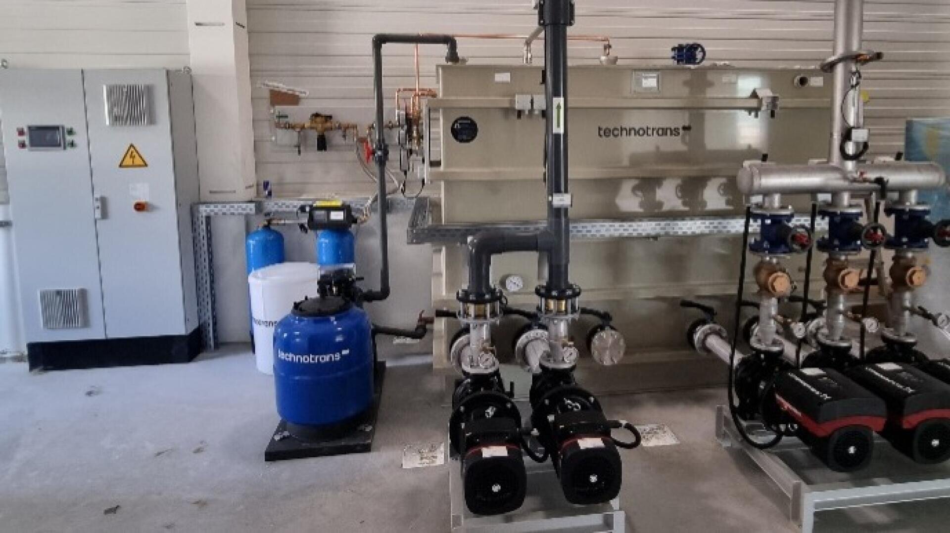 Eine Wasseraufbereitungsanlage in einem industriellen Umfeld mit Tanks mit der Aufschrift technotrans, Schalttafeln, Rohren, Pumpen und Ventilen, die alle vor einer weißen Wand angeordnet sind.