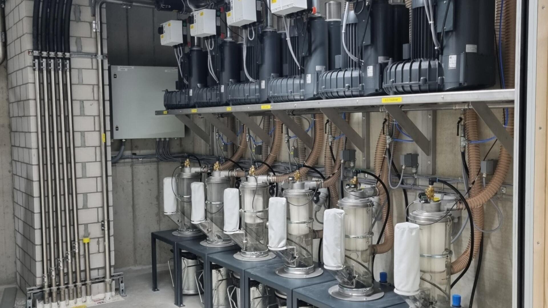 Eine Reihe von industriellen Vakuumpumpen und -filtern, die mit Schläuchen verbunden sind, steht auf Metallregalen in einem betonierten Technikraum mit freiliegenden Rohren und elektrischen Kabeln.