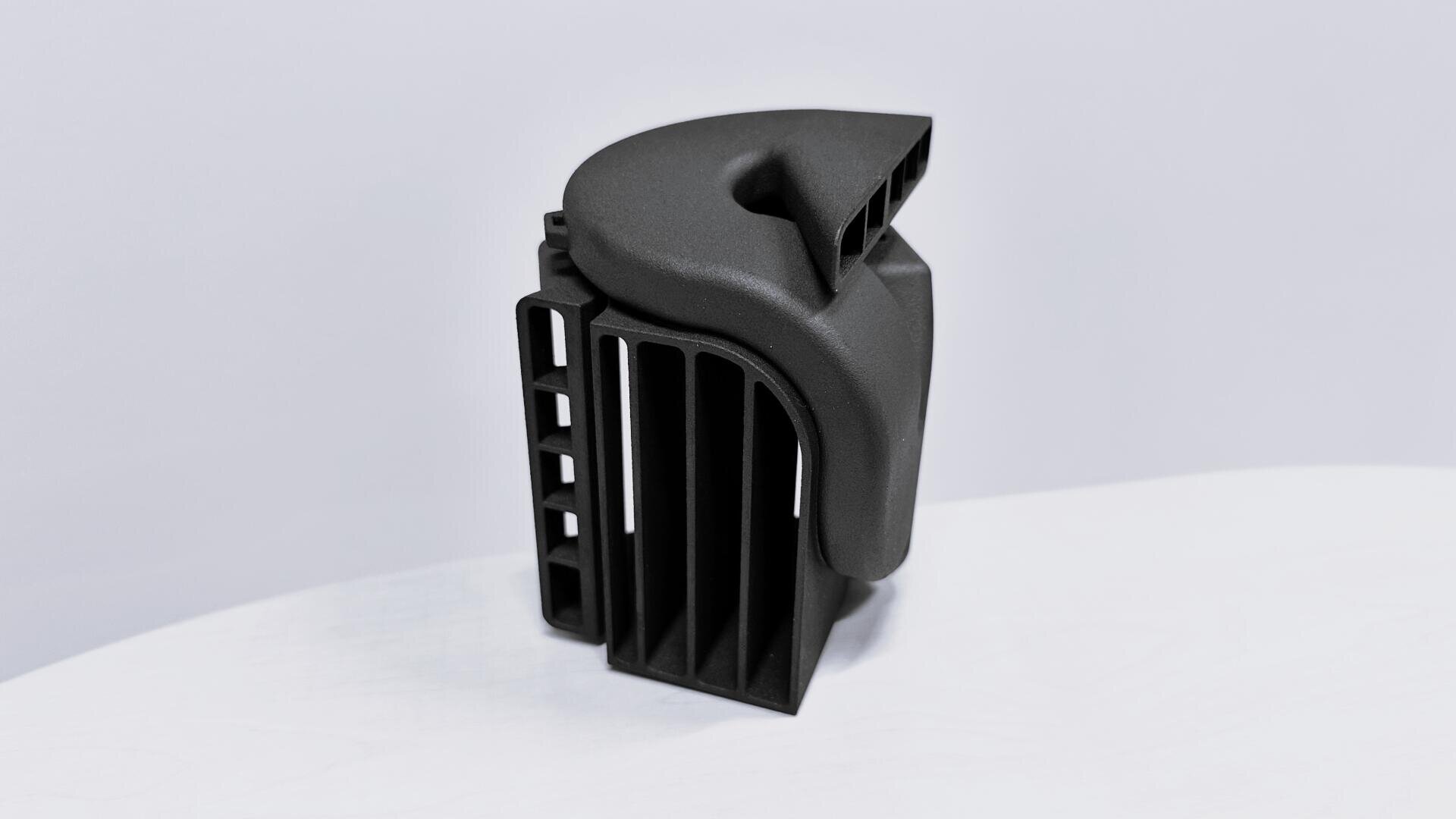 Ein schwarzes, im 3D-Druckverfahren hergestelltes Kunststoffteil mit Lüftungsschlitzen und gewölbten Oberflächen steht aufrecht auf einer weißen Fläche vor einem hellgrauen Hintergrund.