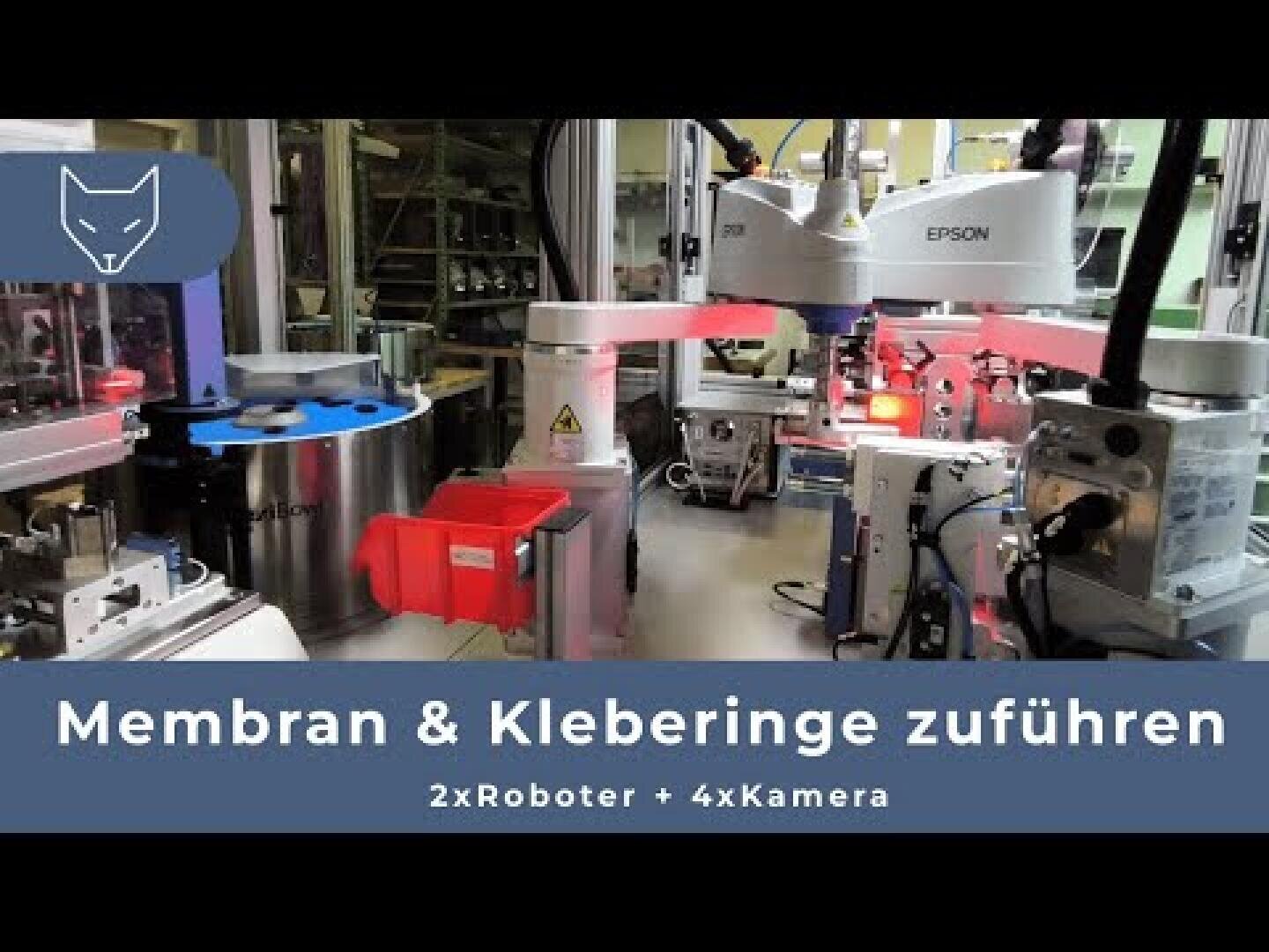 Eine Roboter-Montagelinie mit zwei Epson-Robotern und mehreren Kameras, die Bauteile verarbeiten. Der Text auf Deutsch lautet: Membran & Kleberinge zuführen, 2xRoboter + 4xKamera. In der oberen linken Ecke ist ein Katzenlogo zu sehen.