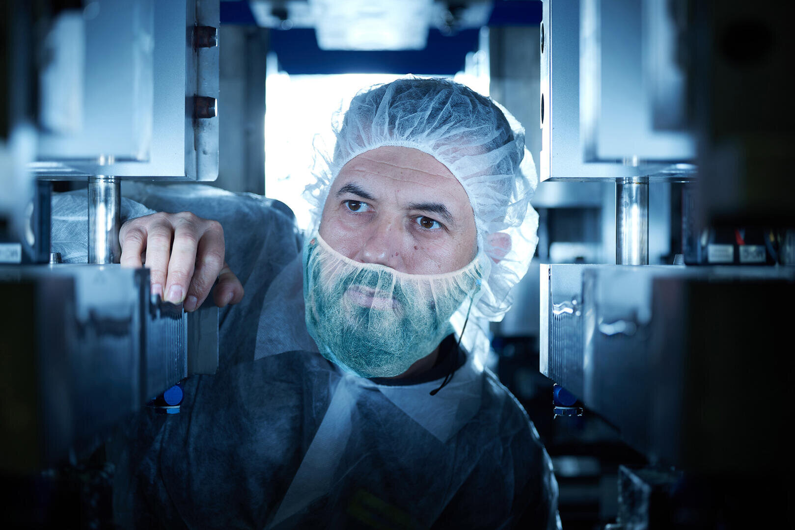Ein Mann, der ein Haarnetz, einen Bartschutz und einen Schutzkittel trägt, inspiziert Maschinen in einer sauberen, sterilen Industrieumgebung, möglicherweise in einem Labor oder einer Produktionsstätte.