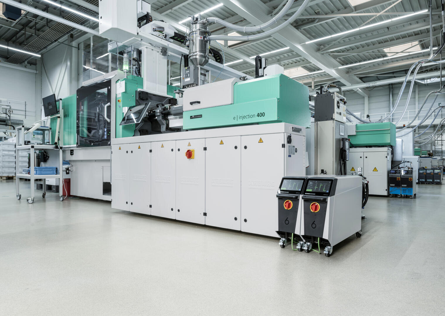 Eine moderne Spritzgießmaschine mit einem Bedienpult steht in einer sauberen, hellen Industriehalle mit hohen Decken, umgeben von anderen Produktionsanlagen.
