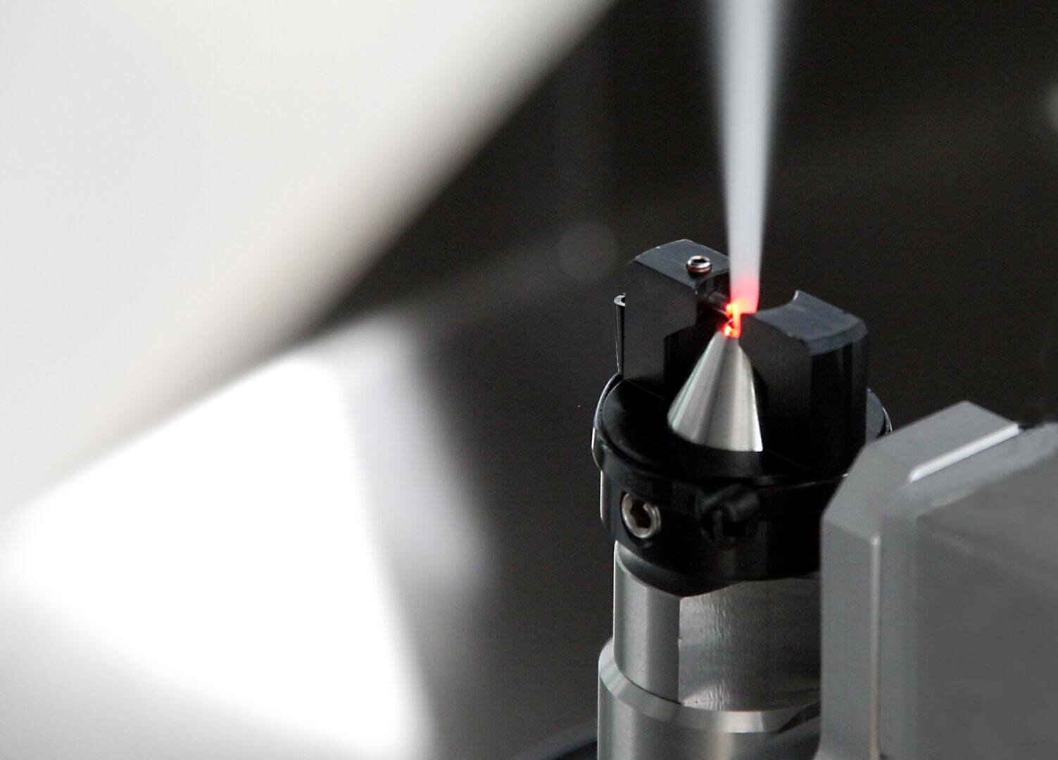 Gros plan sur un instrument scientifique en métal émettant un faisceau de lumière blanche concentré, avec une petite lumière rouge à son extrémité, dans un environnement de laboratoire moderne.