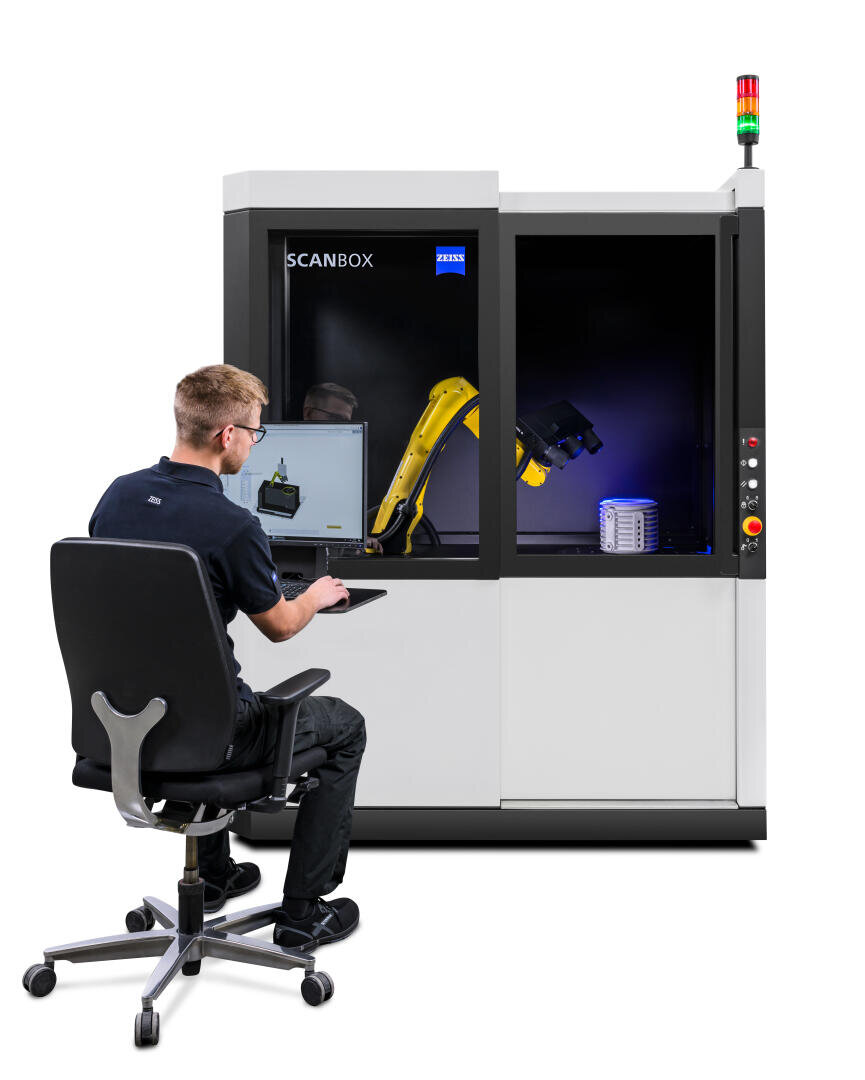 Ein Techniker sitzt an einem Computerarbeitsplatz und bedient eine ZEISS ScanBox, ein automatisches 3D-Scansystem mit einem Roboterarm und einem Metallobjekt im Inneren, das für die industrielle Inspektion und Qualitätskontrolle verwendet wird.