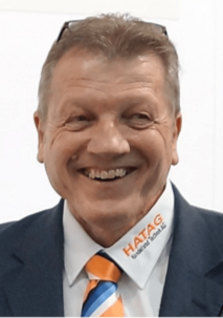 Ein lächelnder Mann mittleren Alters mit kurzen hellbraunen Haaren, der einen Anzug, eine gestreifte orange-blaue Krawatte und ein weißes Hemd mit der Aufschrift HATAG und WORK SHOP am Kragen trägt. Auf seinem Kopf ruht eine Brille.