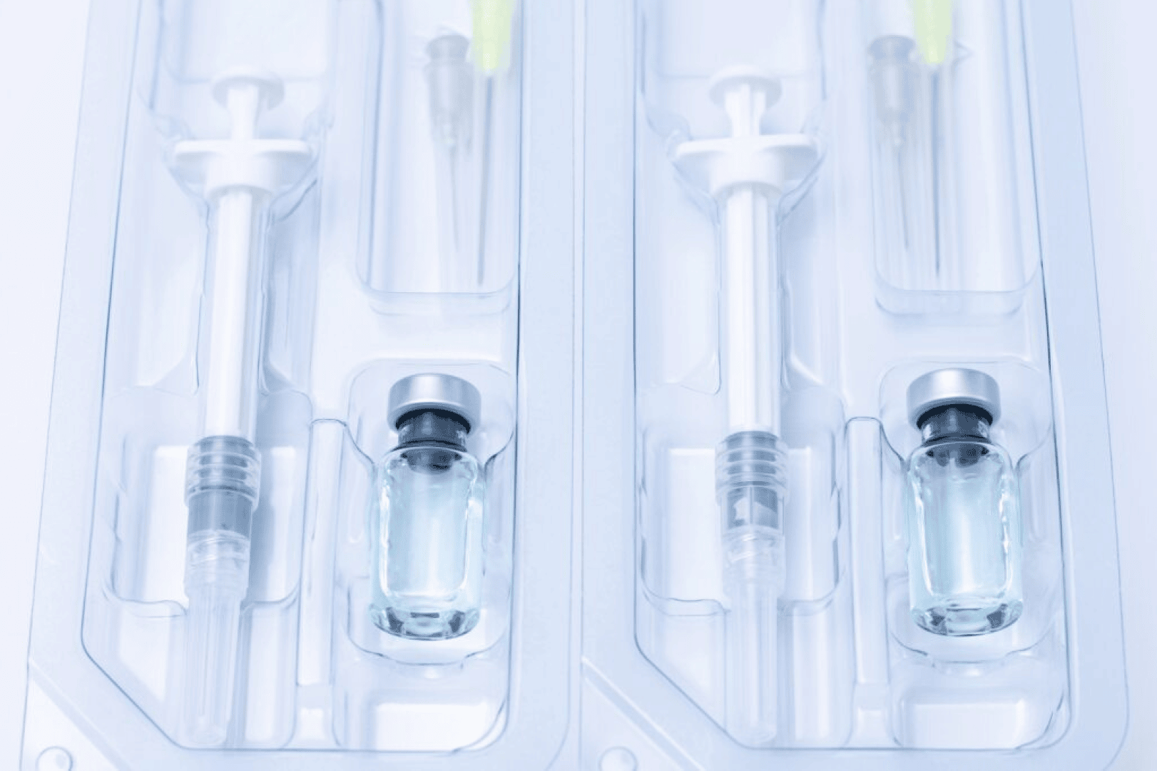 Zwei versiegelte Verpackungen mit je einer Spritze und einem kleinen Glasfläschchen, gefüllt mit einer klaren Flüssigkeit, möglicherweise ein Medikament oder ein Impfstoff, vor einem hellen Hintergrund.