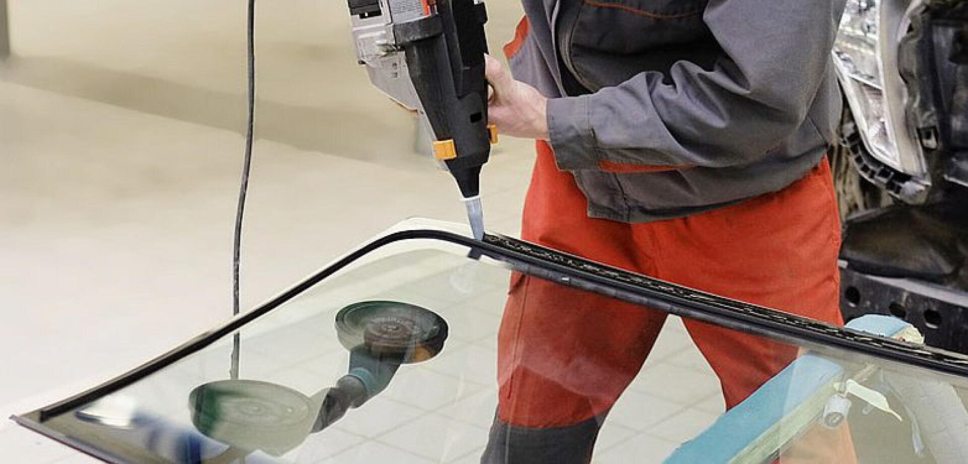 Une personne en uniforme rouge et gris utilise un outil électrique pour appliquer de l'adhésif sur le bord d'un pare-brise de voiture, afin de le préparer à l'installation dans un atelier de réparation automobile.