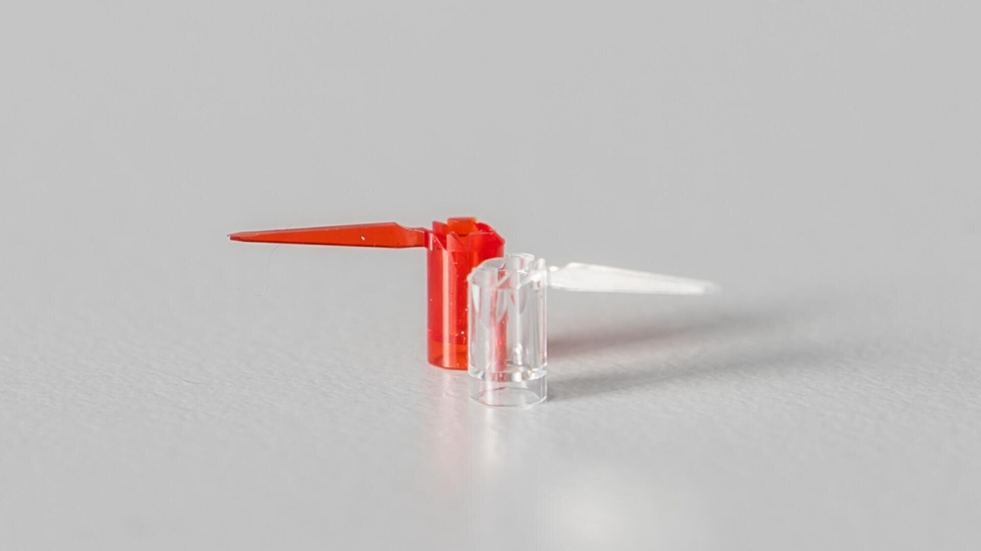 Eine kleine Plastikspielzeugfigur mit einem roten und einem durchsichtigen Flügel steht aufrecht auf einer glatten, hellgrauen Oberfläche. Die Figur wirft einen schwachen Schatten.