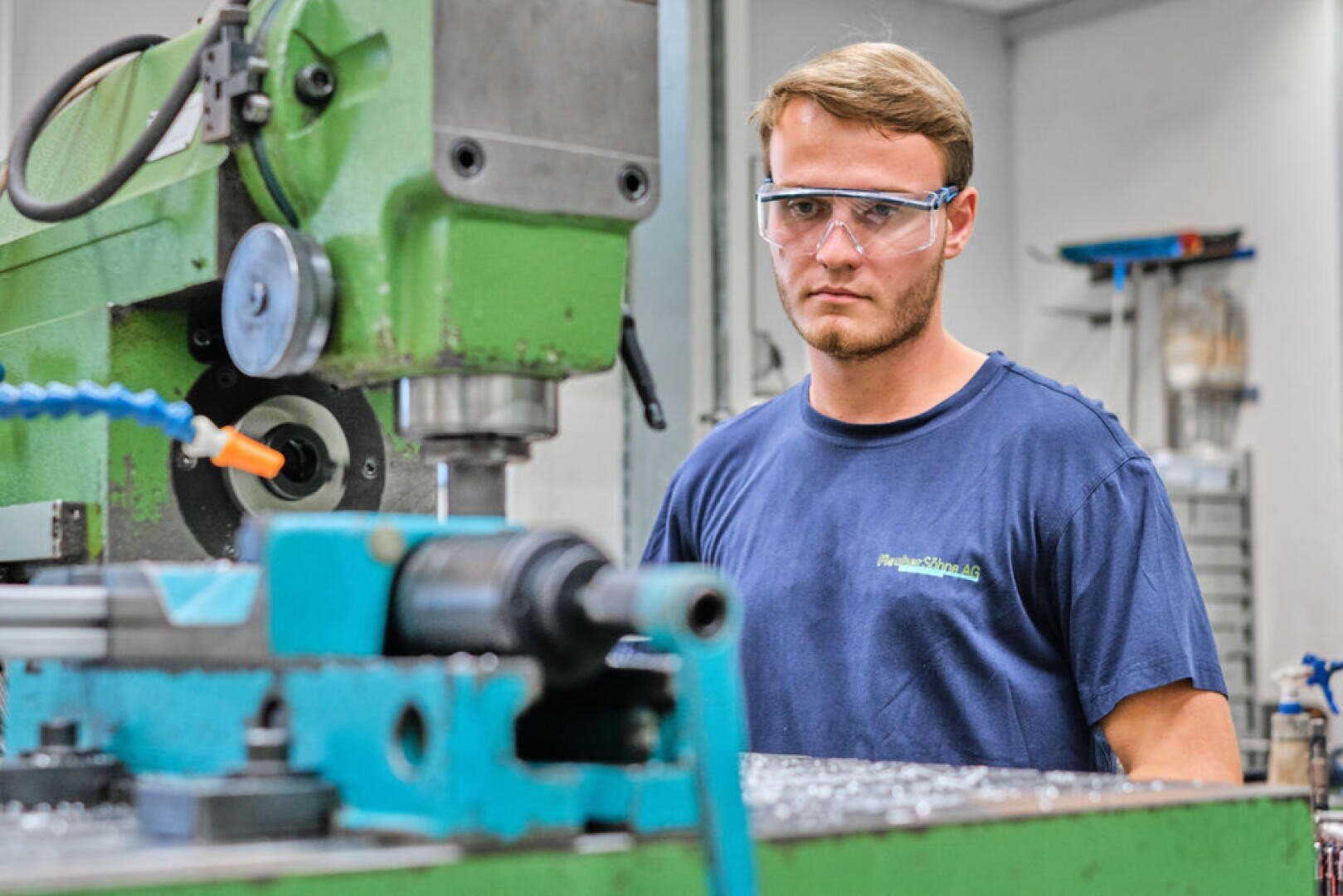 Un homme portant des lunettes de protection et une chemise bleue fait fonctionner une machine industrielle verte dans un atelier et se concentre sur son travail. Des copeaux de métal sont visibles sur le sol à côté de la machine.