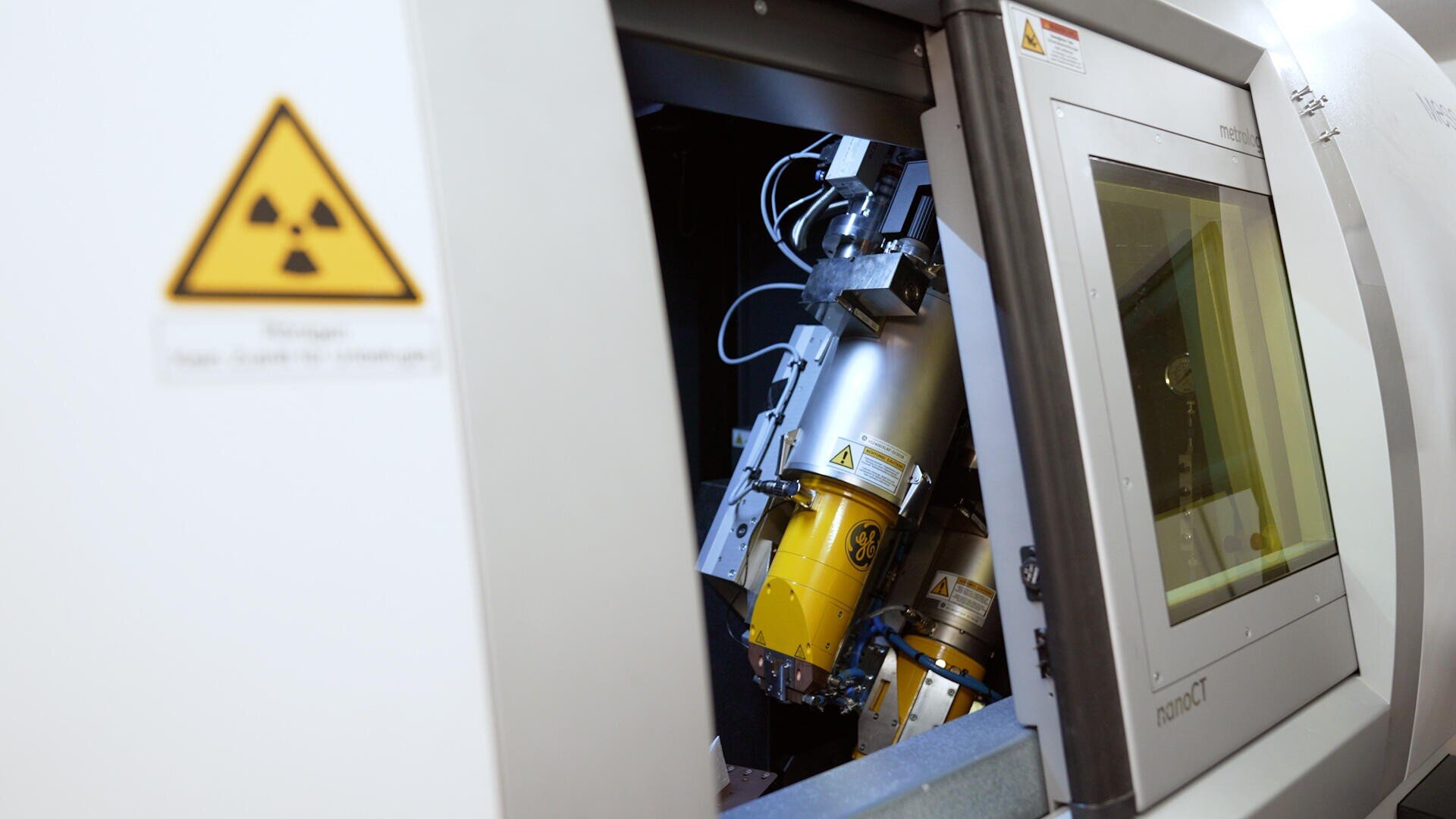 Nahaufnahme einer Industrieanlage im Inneren einer Maschine. Links ist ein Strahlungswarnschild zu sehen, im geöffneten Bedienfeld sind mechanische Komponenten und Verkabelung zu sehen. Rechts sind ein Monitor und ein Bedienfeld zu sehen.