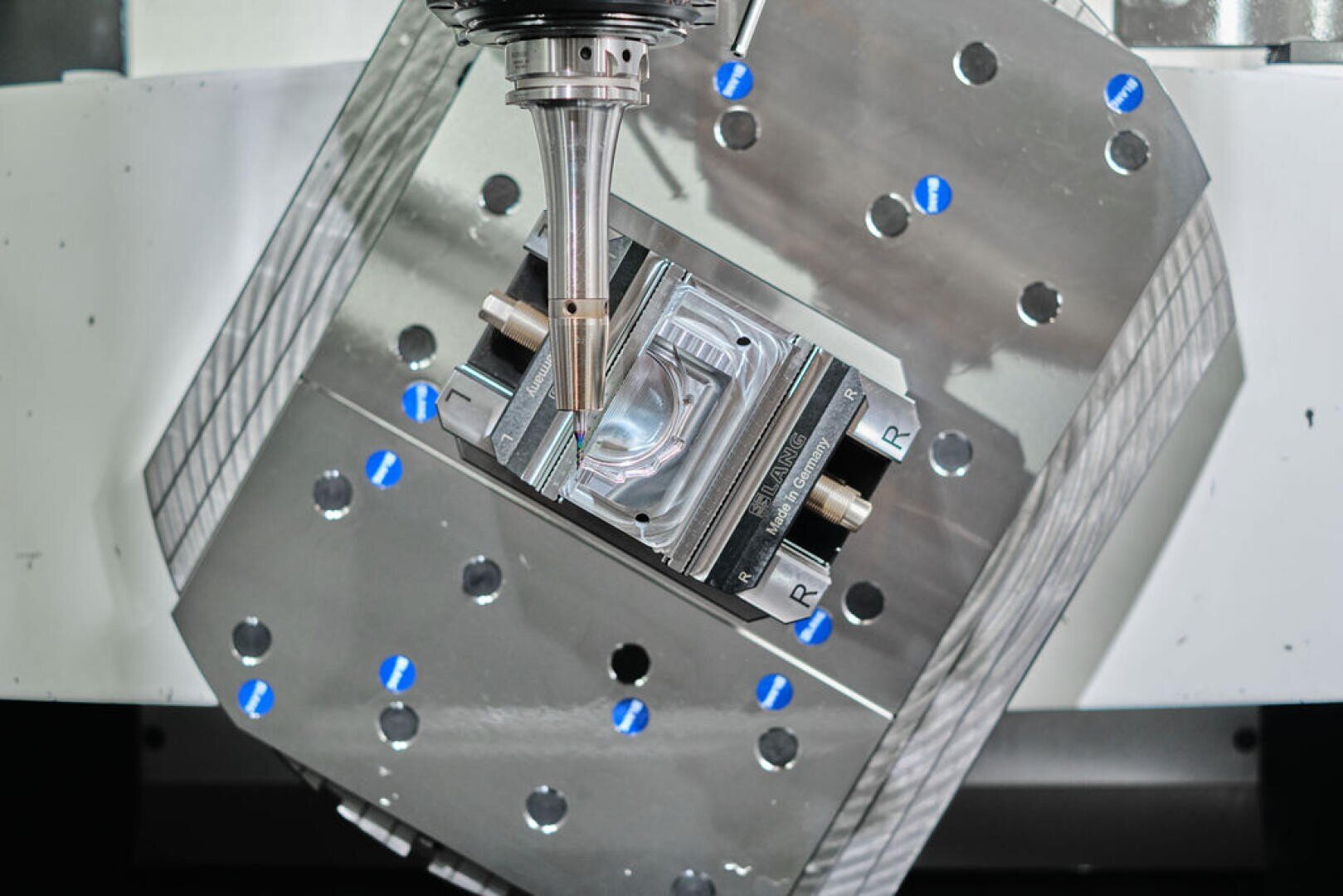 Une tête de machine-outil à commande numérique usine une pièce métallique fixée sur un plateau rotatif afin d'usiner un composant précis et anguleux dans un environnement industriel. Des points bleus et des marques gravées sont visibles sur les surfaces métalliques.