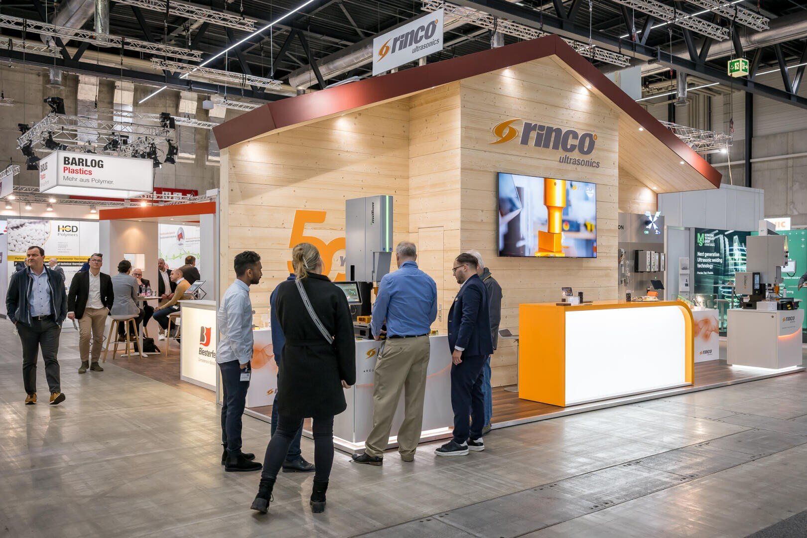 Un stand de salon pour Rinco Ultrasonics, comprenant une structure en bois avec des présentoirs et de l'équipement. Plusieurs personnes interagissent sur le stand, tandis que d'autres passent dans le hall d'exposition.
