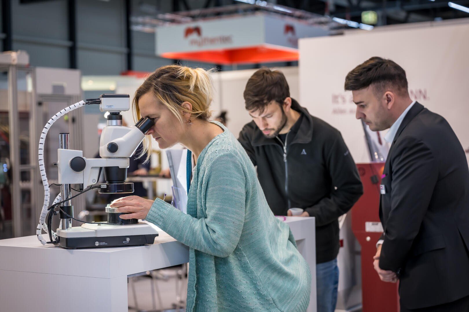 Une femme regarde dans un microscope, tandis que deux hommes observent une exposition de sciences et de techniques à proximité. L'environnement semble professionnel, avec des stands et des appareils en arrière-plan.