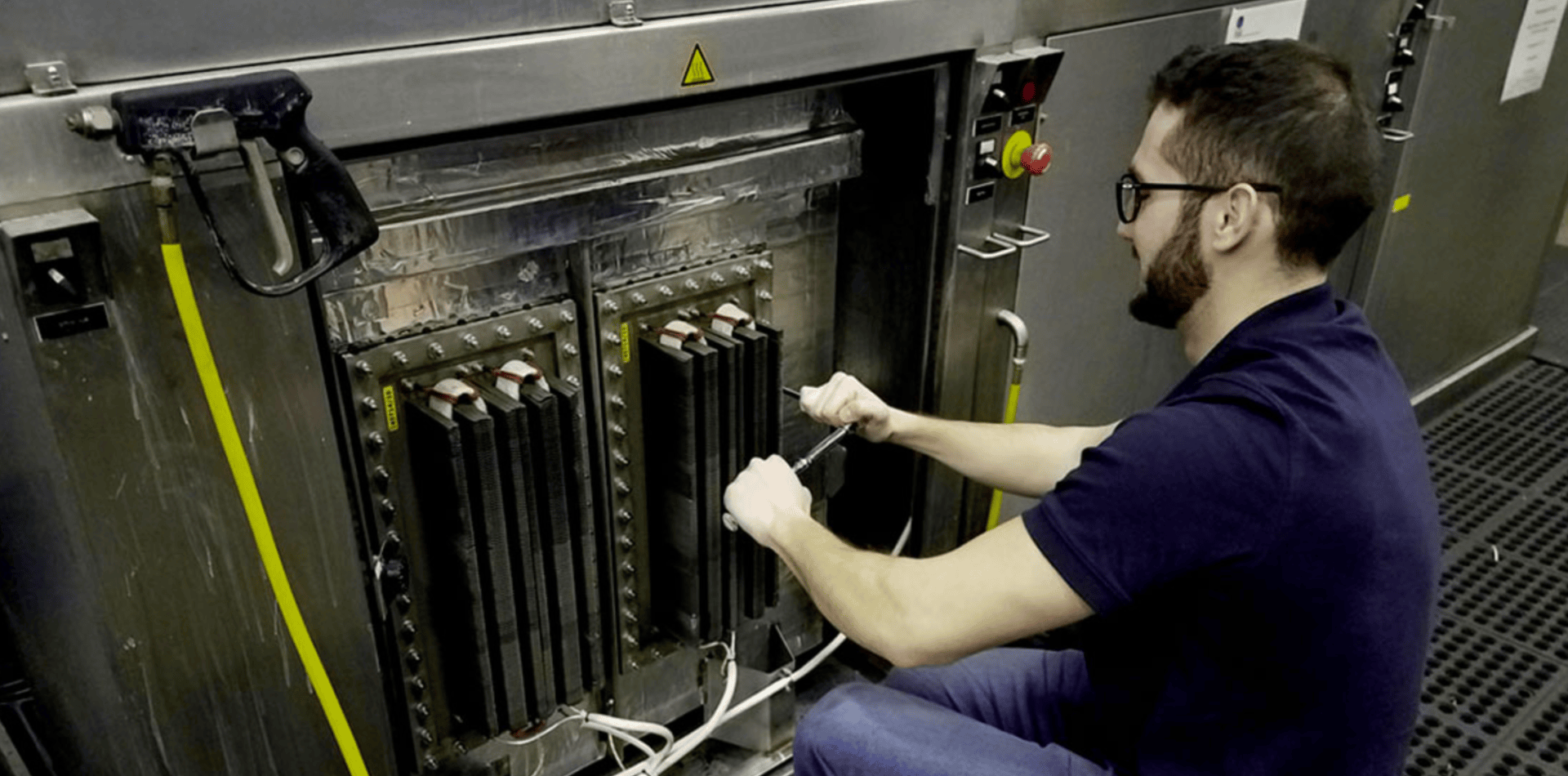 Un homme portant des lunettes et une chemise marine est agenouillé devant un équipement industriel, ajustant des câbles reliés à deux grands panneaux noirs verticaux à l'intérieur d'une chambre métallique.