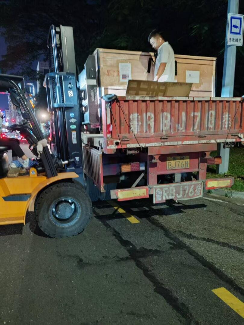 Ein Mann steht auf einer roten LKW-Ladefläche und führt eine große Holzkiste, die von einem gelben Gabelstapler auf einer nächtlichen Straße angehoben wird. Das Nummernschild des Lastwagens ist sichtbar, und im Hintergrund sind Straßenschilder und Verkehr zu sehen.