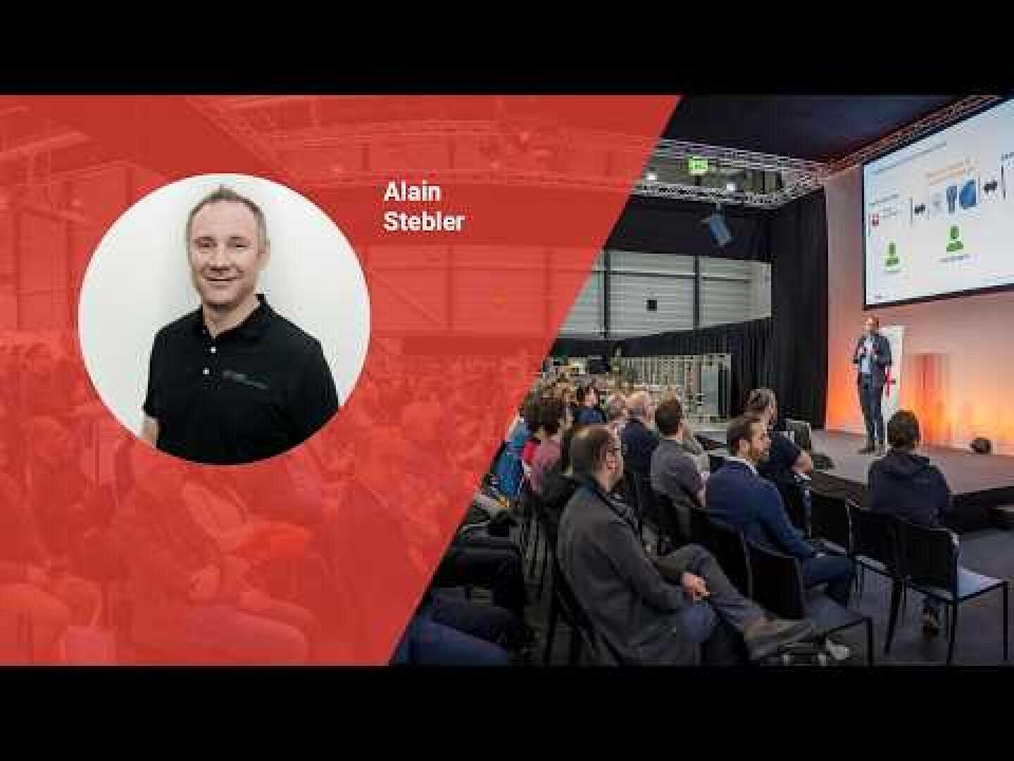 Präsentationsszene mit links dem Foto und dem Namen von Alain Stebler auf rotem Hintergrund; rechts spricht ein Mann auf der Bühne zu einem Publikum, das in einem modernen Konferenzraum sitzt, wobei hinter ihm ein Dia projiziert wird.