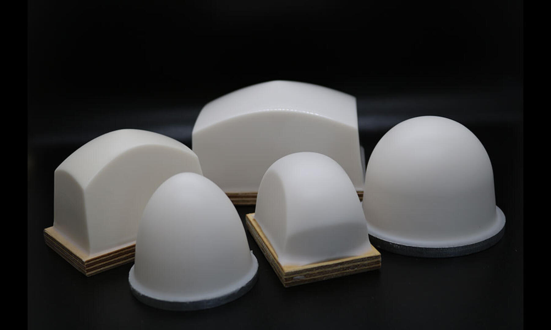 Cinq objets lisses, blancs, en forme de dôme ou d'arche, avec une base en bois ou foncée, sont disposés sur un fond noir. Trois sont en forme de dôme et deux ont un sommet bombé et des côtés plats.