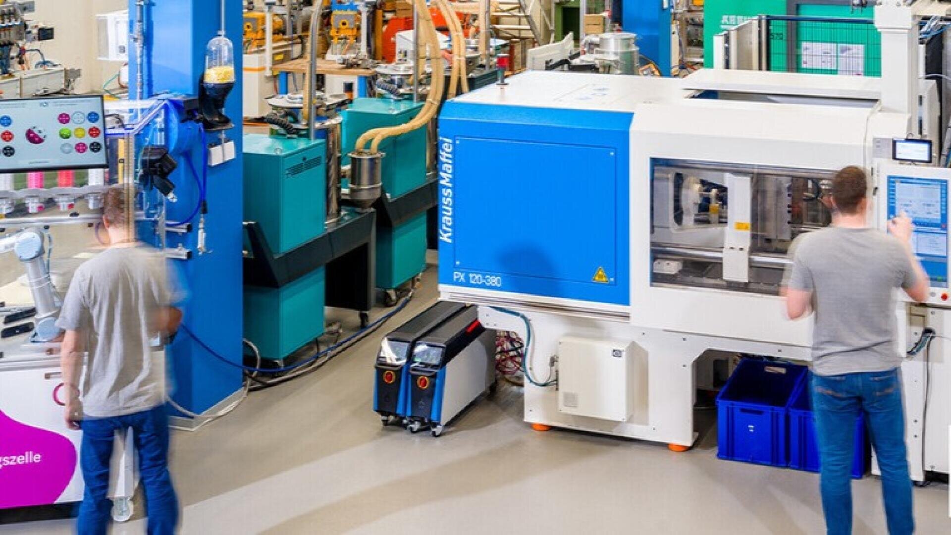 Deux personnes travaillent dans un hall industriel bien éclairé avec des machines de moulage par injection, dont une grande installation KraussMaffei bleue et blanche, entourée d'autres équipements et de conteneurs de stockage.
