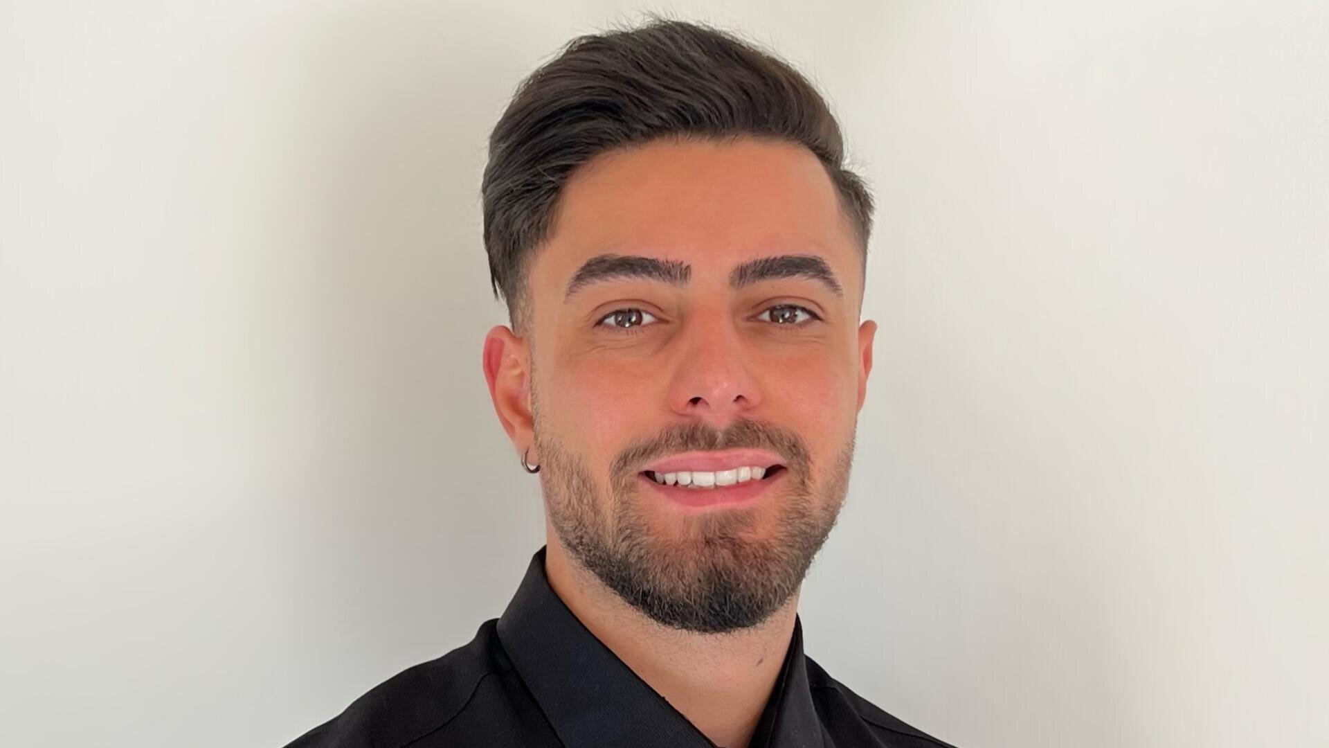 Ein Mann mit kurzen dunklen Haaren und gestutztem Bart, der ein schwarzes Button-up-Hemd trägt, steht vor einem schlichten weißen Hintergrund und lächelt in die Kamera.