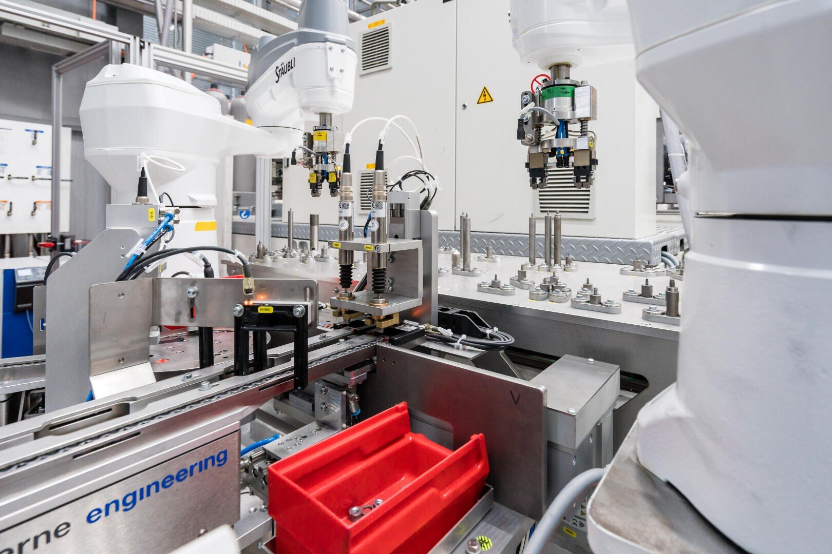 Des robots industriels automatisés effectuent des tâches d'assemblage dans une usine moderne. La scène montre des bras de robots, des tapis roulants, des fils et des machines avec un conteneur en plastique rouge au premier plan.