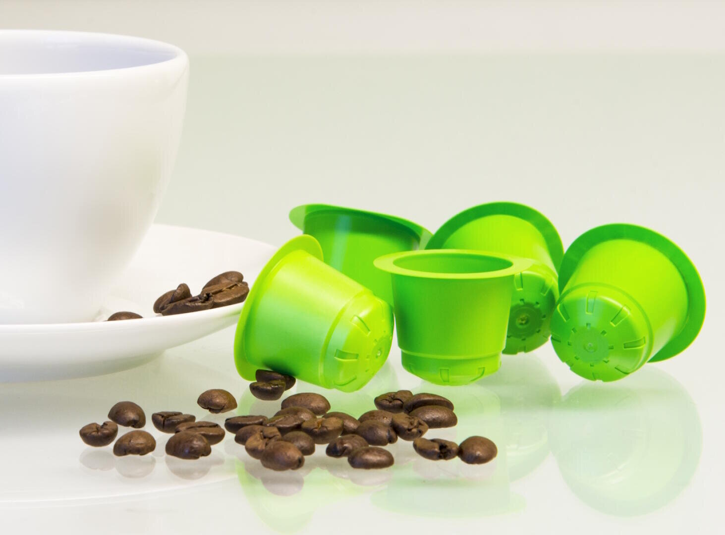 Vier grüne Kaffeekapseln und Kaffeebohnen sind auf einer weissen Fläche neben einer weissen Tasse und Untertasse verstreut. Die Szene ist hell beleuchtet, was ihr ein frisches und sauberes Aussehen verleiht.