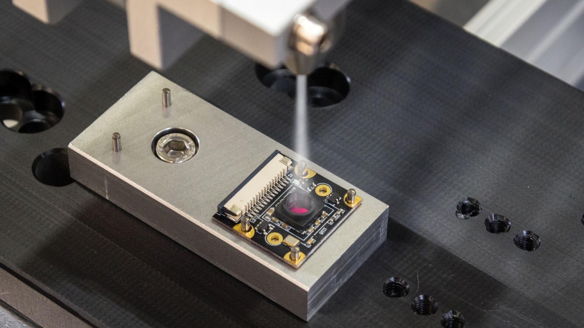 Gros plan sur un petit composant électronique pulvérisé ou revêtu avec précision par une buse de machine, monté sur un support métallique avec des vis et une surface de travail perforée de couleur sombre.