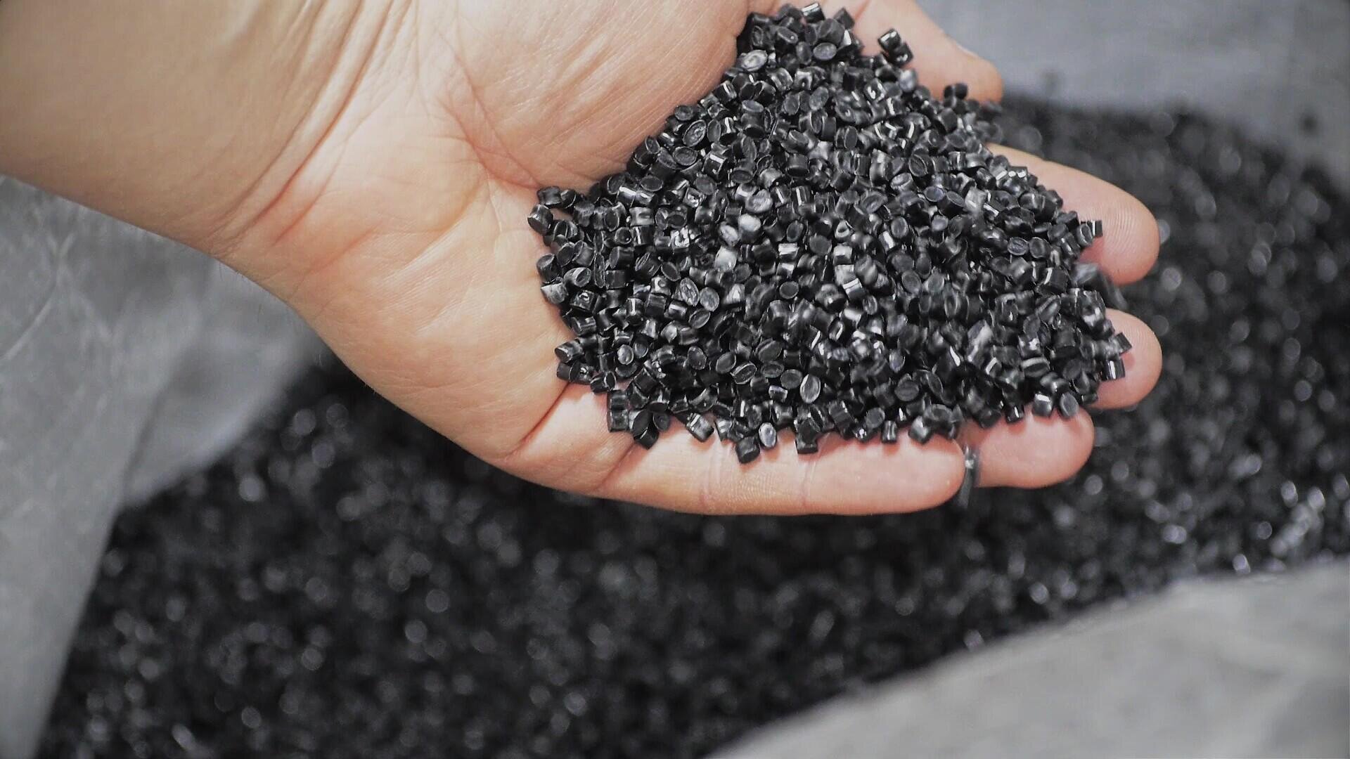 Eine Hand hält einen Haufen kleiner schwarzer Plastikkügelchen, im Hintergrund sind weitere Kügelchen zu sehen.