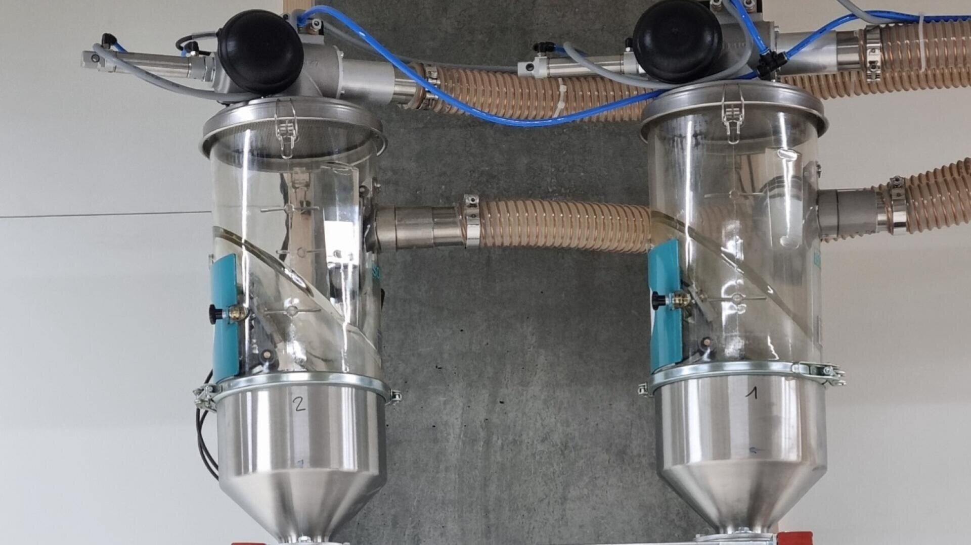 Zwei industrielle Vakuumtrichter aus Glas und Metall mit angeschlossenen Schläuchen und nummerierten Etiketten sind an einer Wand montiert und werden für den Transport und die Dosierung von Granulat oder Pulvern verwendet.
