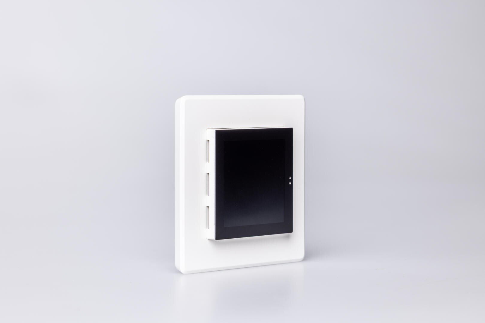 Ein weißes, an der Wand montiertes Smart Home-Bedienfeld mit schwarzem Touchscreen-Display vor einem schlichten hellgrauen Hintergrund.