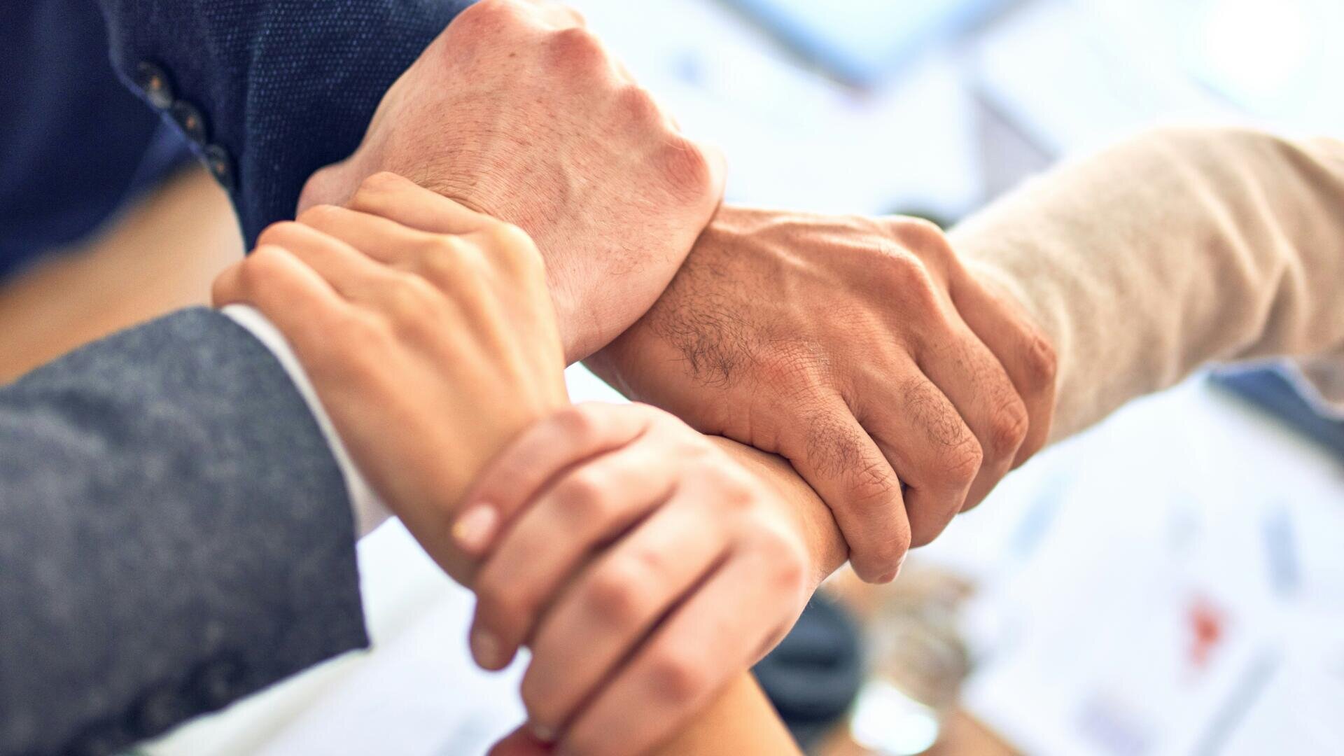 Quatre personnes se tiennent par le poignet dans un carré, symbolisant le travail d'équipe, l'unité et le soutien, avec des tons de peau et des vêtements différents. L'arrière-plan est flou et semble être un bureau.