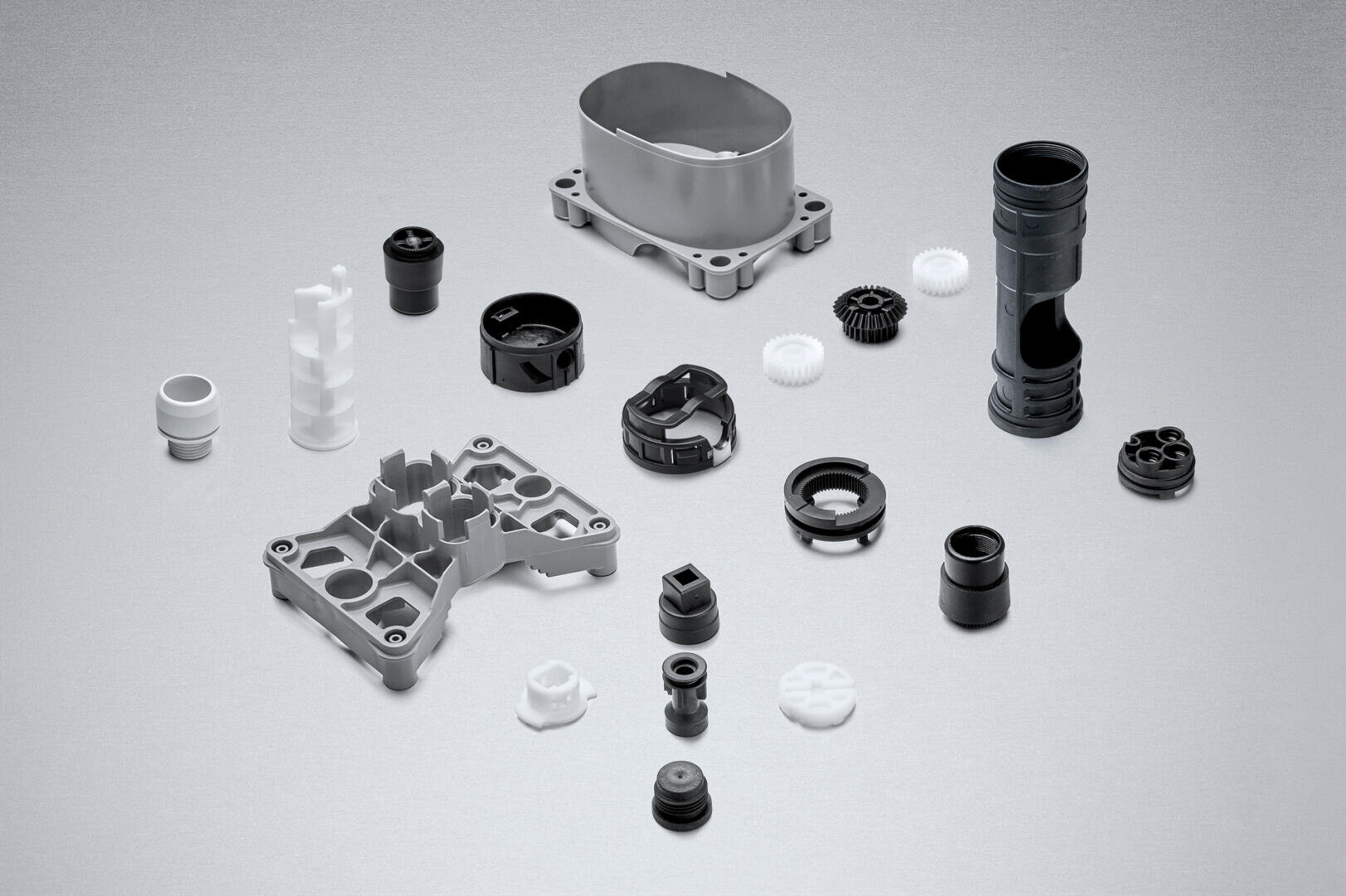 Eine Vielzahl schwarzer, weißer und grauer mechanischer Teile und Zahnräder aus Kunststoff und Metall sind übersichtlich auf einer einfarbig grauen Fläche angeordnet.
