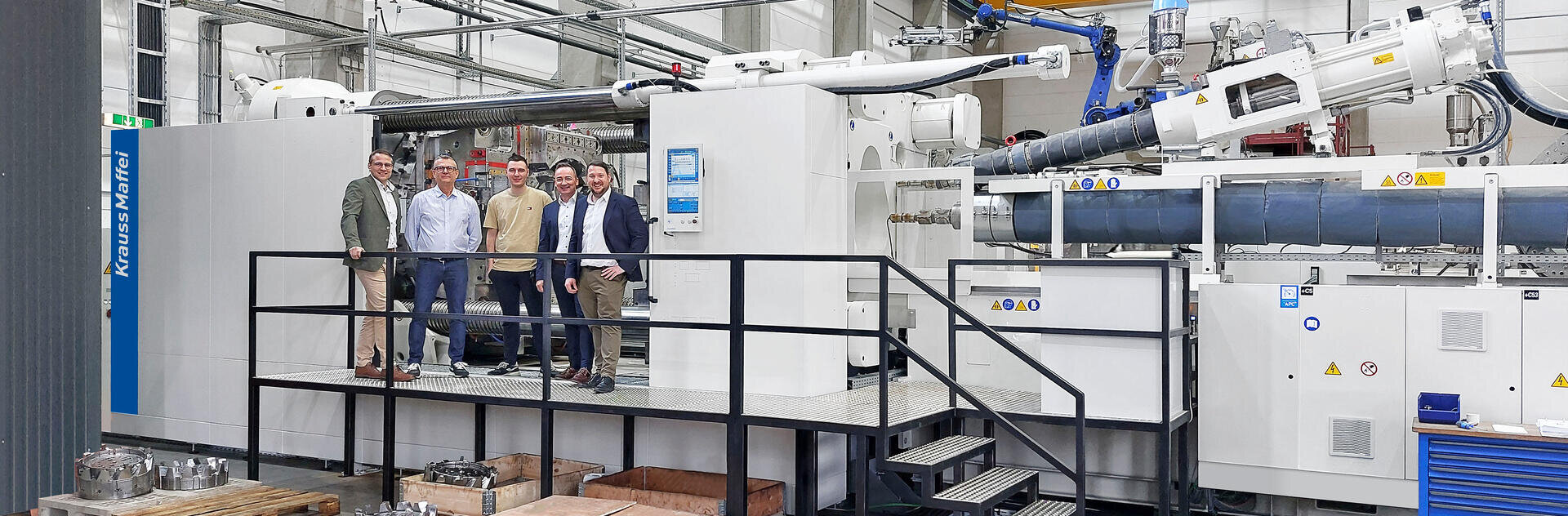 Vier Männer in Geschäftskleidung stehen auf einem Podest vor einer großen industriellen Spritzgießmaschine in einer Fabrik, mit Rohren und Maschinen im Hintergrund.