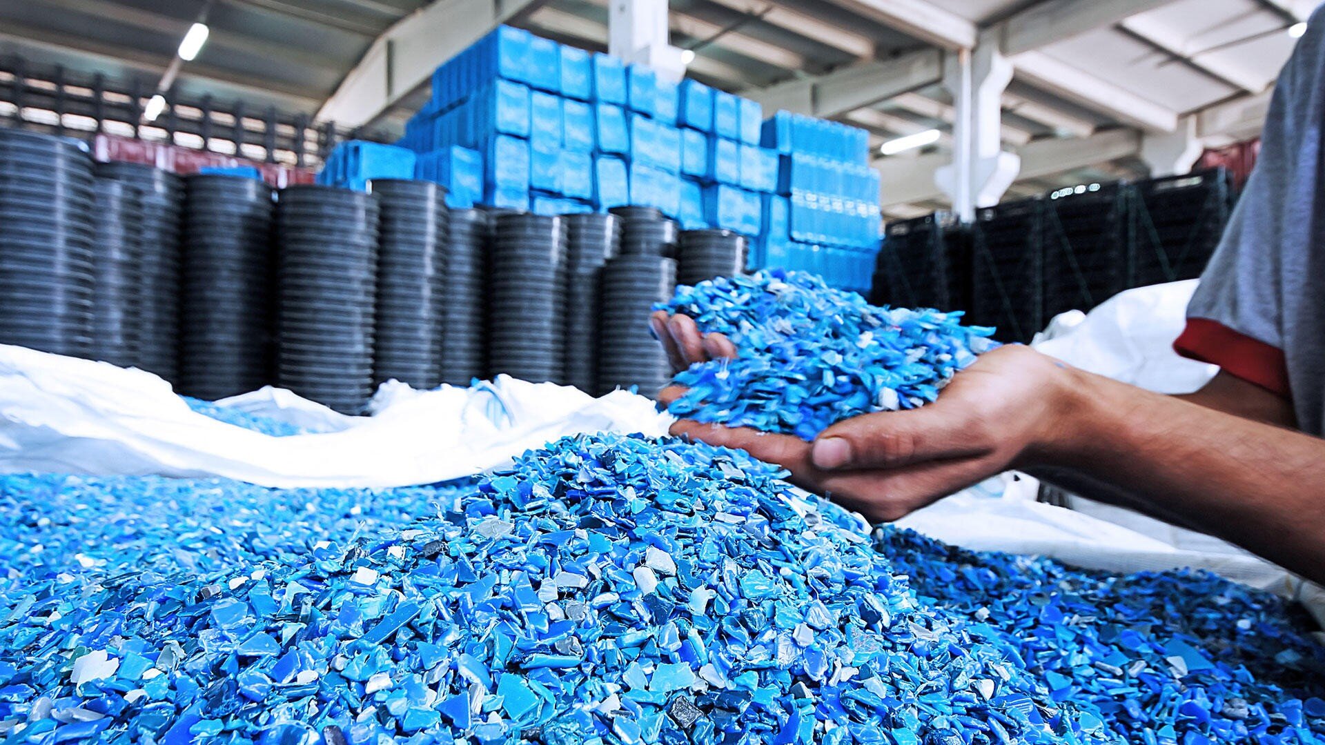 Eine Person hält eine Handvoll blauer Plastikfragmente in einer Fabrik, mit einem großen Haufen von Plastikteilen im Vordergrund und Stapeln von Containern im Hintergrund.