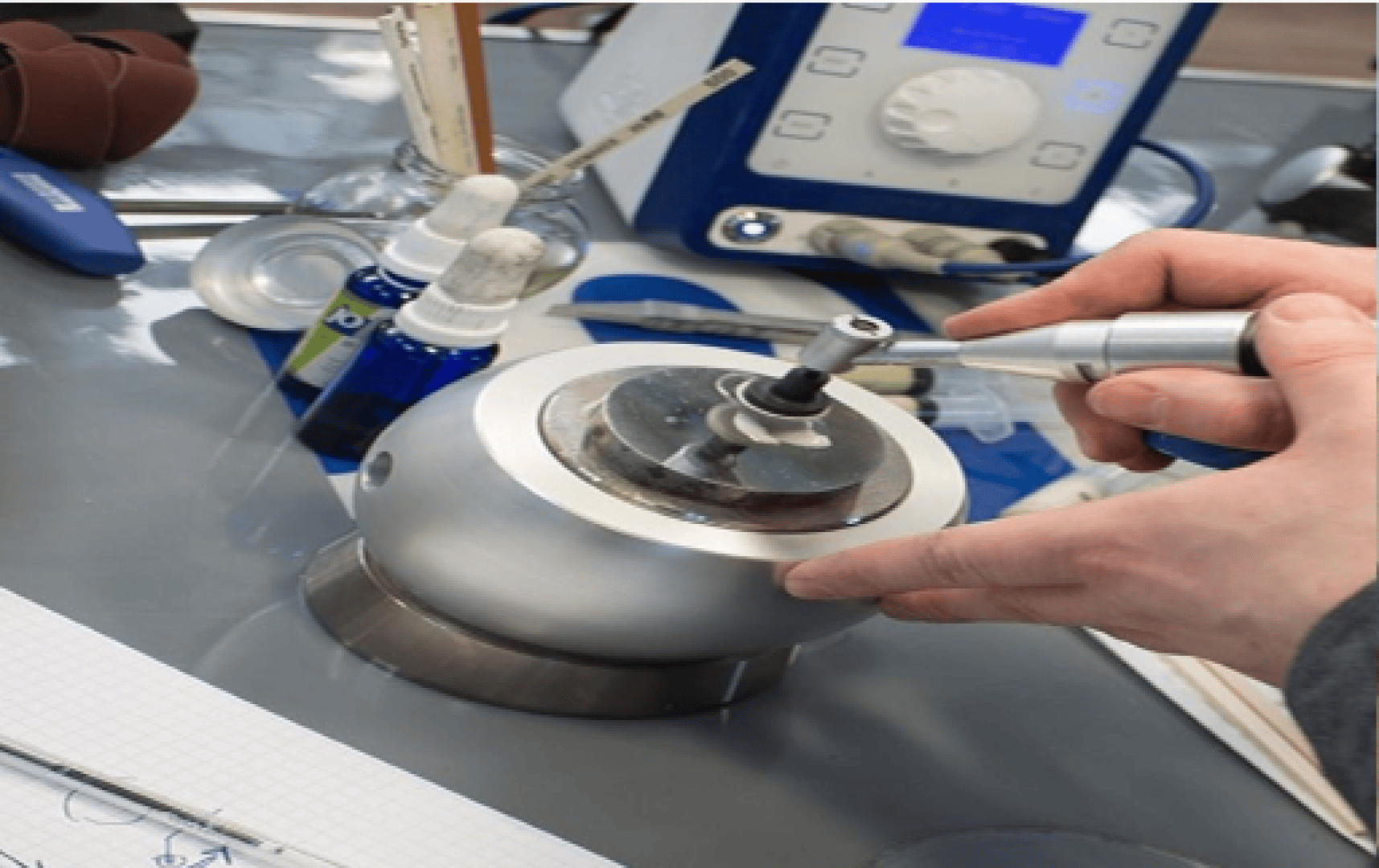 Une personne travaille avec un outil dentaire sur un objet métallique maintenu dans un étau, entourée d'appareils de laboratoire, de bouteilles bleues et d'instruments scientifiques sur un établi.