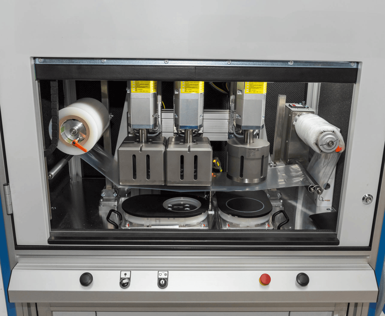 Nahaufnahme einer Industriemaschine mit Rollen, Spulen und automatischen Komponenten in einem Metallgehäuse. Drei vertikale Einheiten sind mit Warnschildern versehen, und an der Vorderseite befinden sich Steuertasten.