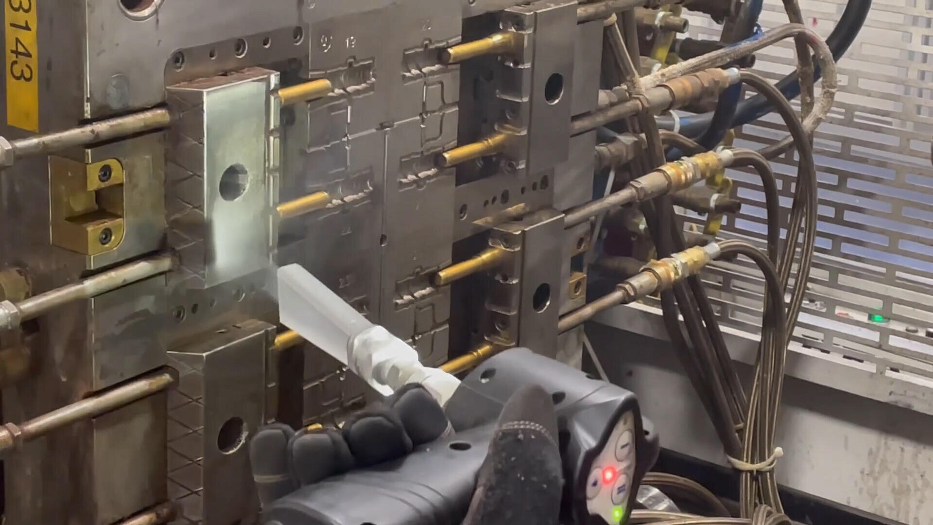 Une main gantée manipule un pistolet à peinture pour appliquer un revêtement ou un agent de démoulage sur un moule à injection métallique auquel sont fixés plusieurs tiges et tuyaux, dans un environnement industriel.