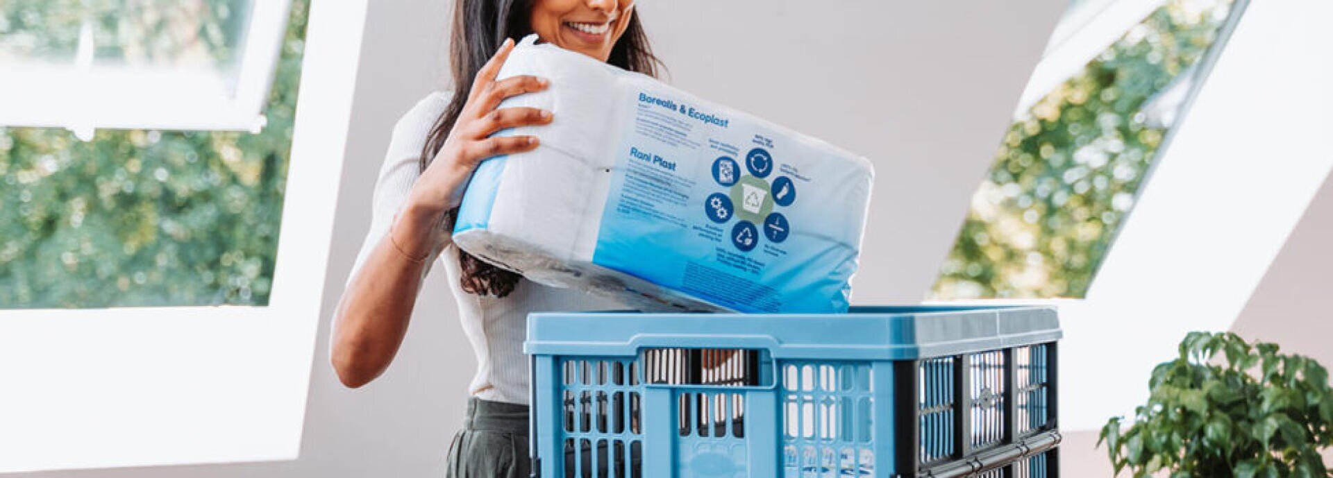 Une femme sourit tout en plaçant un paquet de papier toilette dans un panier à linge bleu à l'intérieur, la lumière du soleil traversant de grandes fenêtres à l'arrière-plan.