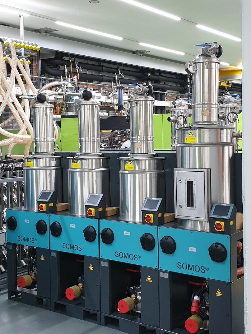 Vier Industriemaschinen aus rostfreiem Stahl mit digitalen Anzeigen und der Aufschrift SOMOS sind in einer Fabrikhalle aufgereiht, umgeben von verschiedenen Rohren, Schläuchen und mechanischen Geräten.