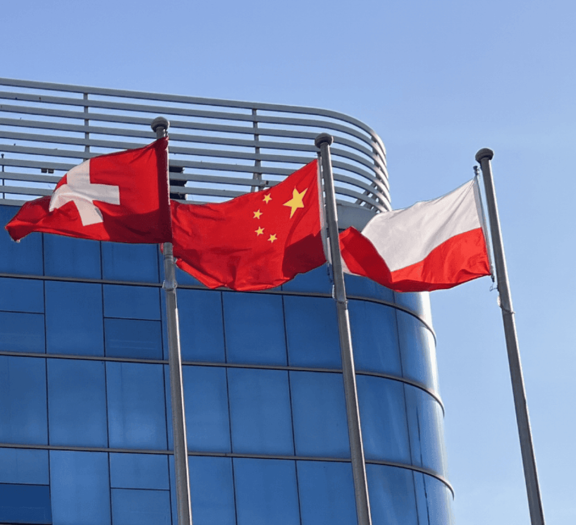 Drei Flaggen - die der Schweiz, Chinas und Polens - wehen an Fahnenmasten vor einem modernen Glasgebäude vor einem strahlend blauen Himmel.