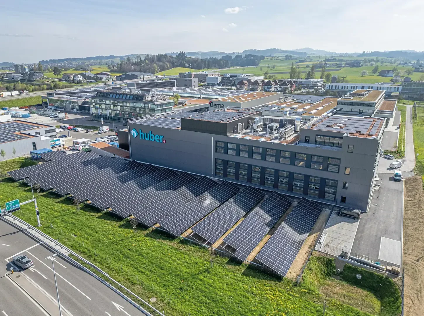 Vue aérienne d'un bâtiment industriel avec le panneau "huber" et une rangée de panneaux solaires installés sur la pelouse devant le bâtiment. D'autres bâtiments industriels, routes et espaces verts se trouvent aux alentours de l'installation.