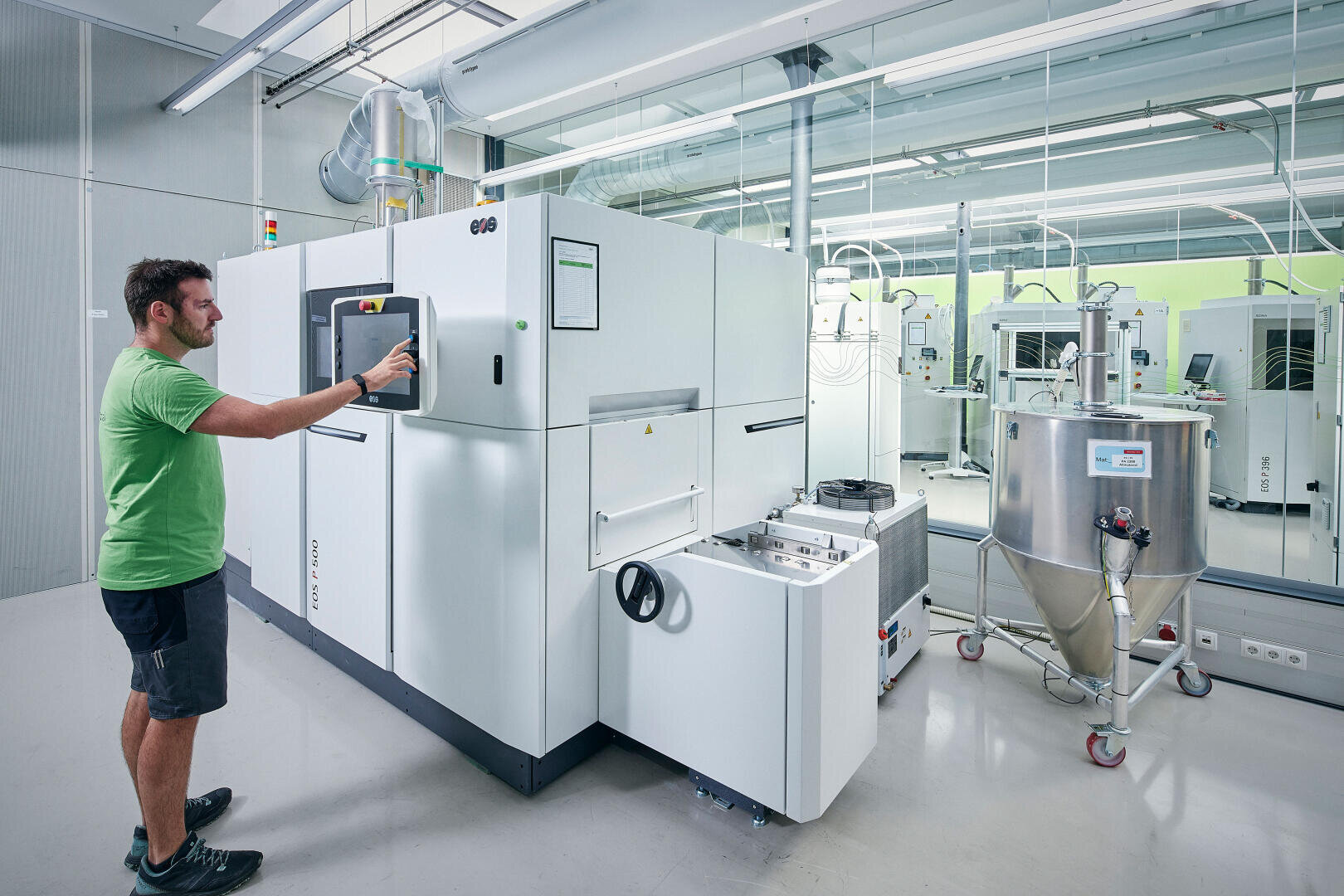 Un homme vêtu d'une chemise verte fait fonctionner une grande imprimante industrielle 3D blanche dans un laboratoire propre et moderne équipé de divers appareils et de murs en verre réfléchissant.