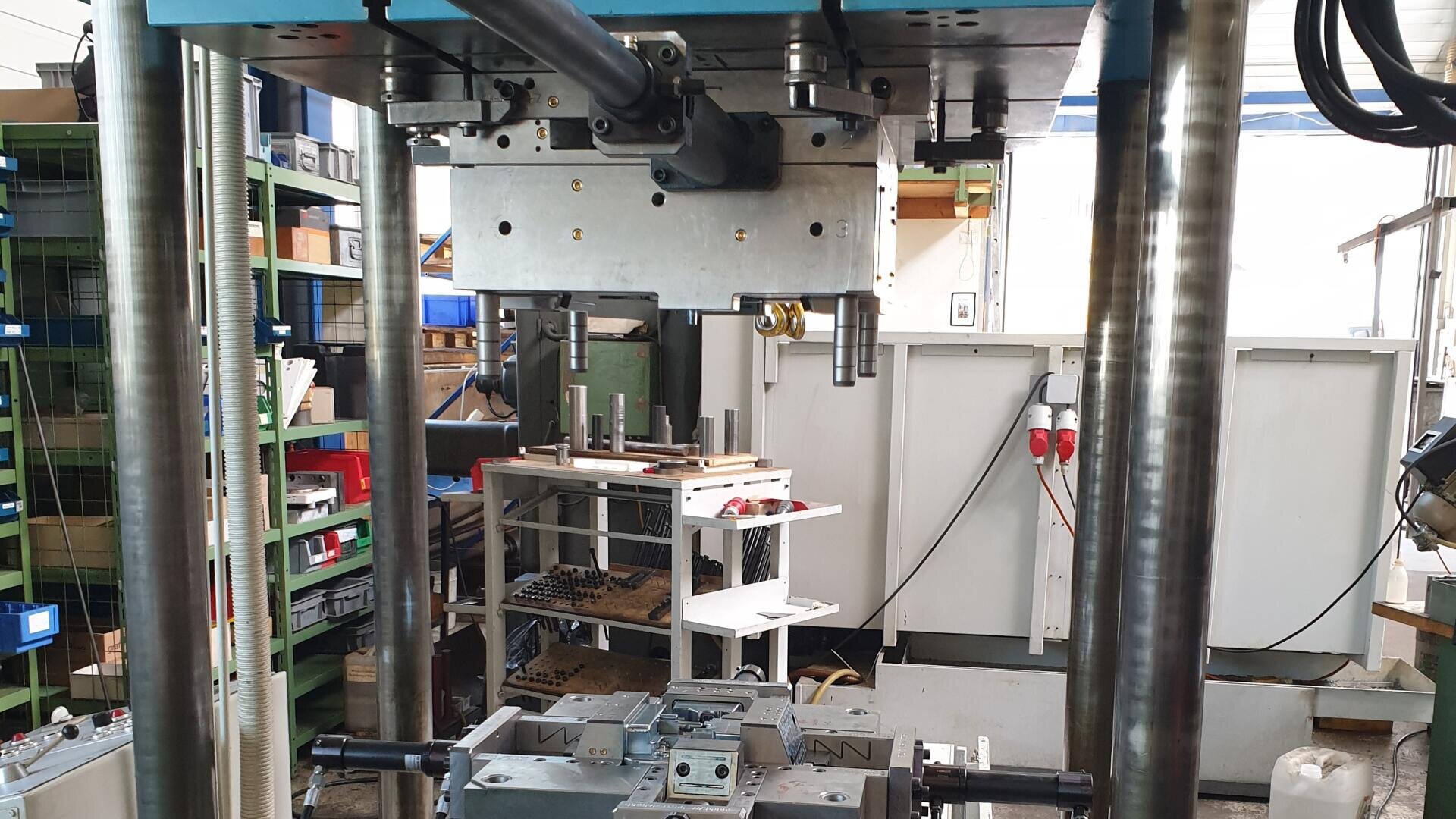 Gros plan sur une machine industrielle dans un atelier, avec en arrière-plan des tiges métalliques, des étagères contenant des outils et du matériel, et des casiers de rangement organisés.